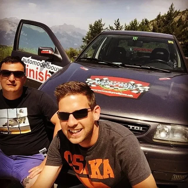 Two men wearing sunglasses smiling in front of a race car with a shirts and a logo on the hood, with mountains and trees in the background.