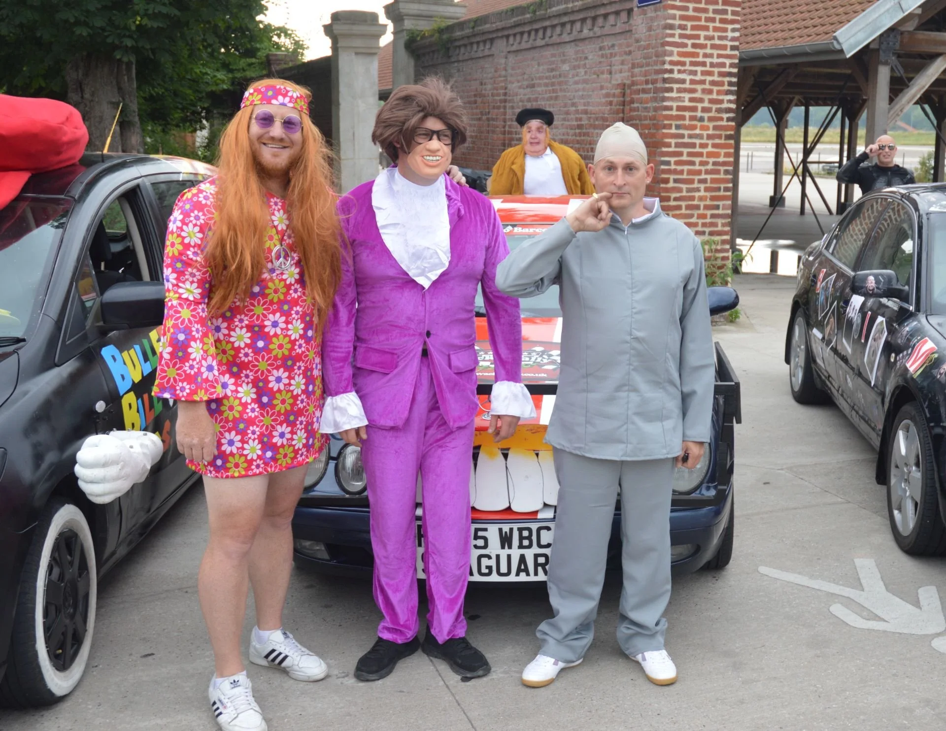 Three individuals dressed in colorful, retro costumes, standing in front of a car with a large painted smiley face on the bumper, during a parade or event, with additional cars and people in the background.