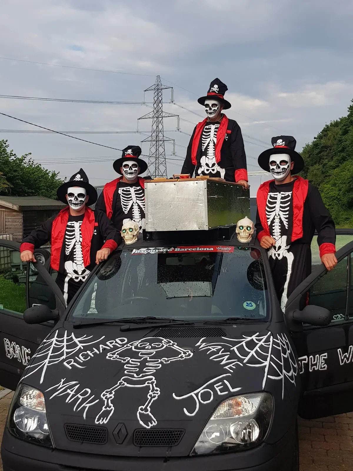 People dressed in skeleton costumes with skull face paint, wearing black hats with skull emblems, standing on and around a decorated black car with a skeleton drawing, cobwebs, and names written on the hood, during daytime.