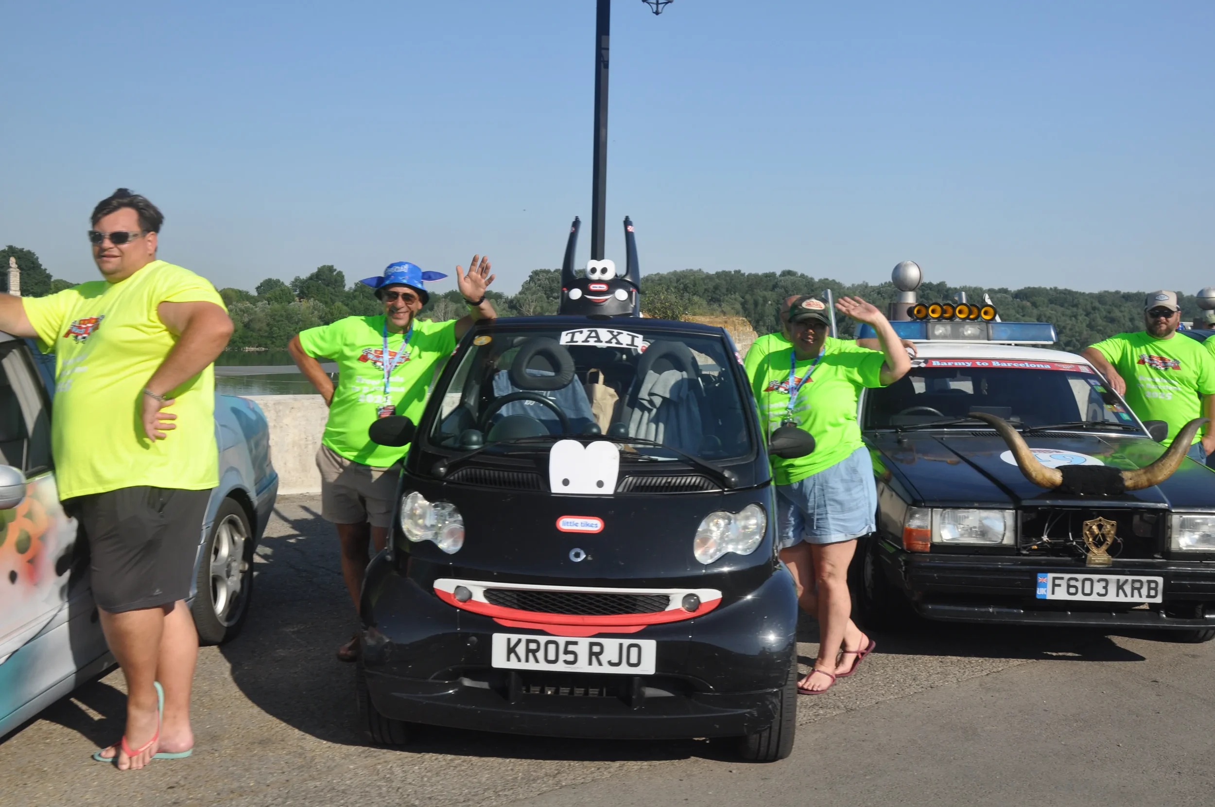 A group of five people wearing neon green shirts, posing outdoors beside decorated cars with humorous and quirky accessories, including a small black electric car with a face sticker, a vintage car with horns, and a mascot on top, on a clear day.