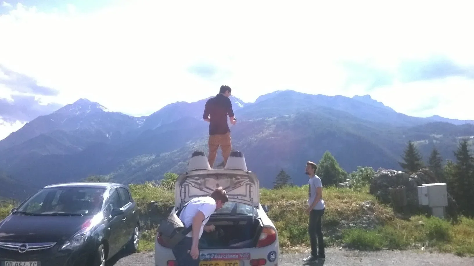Four young men preparing for a trip in a mountainous area with snow-capped peaks in the background. One is standing on the open trunk of a silver car, looking at his phone, another is bent over in the trunk, and two others are standing nearby.