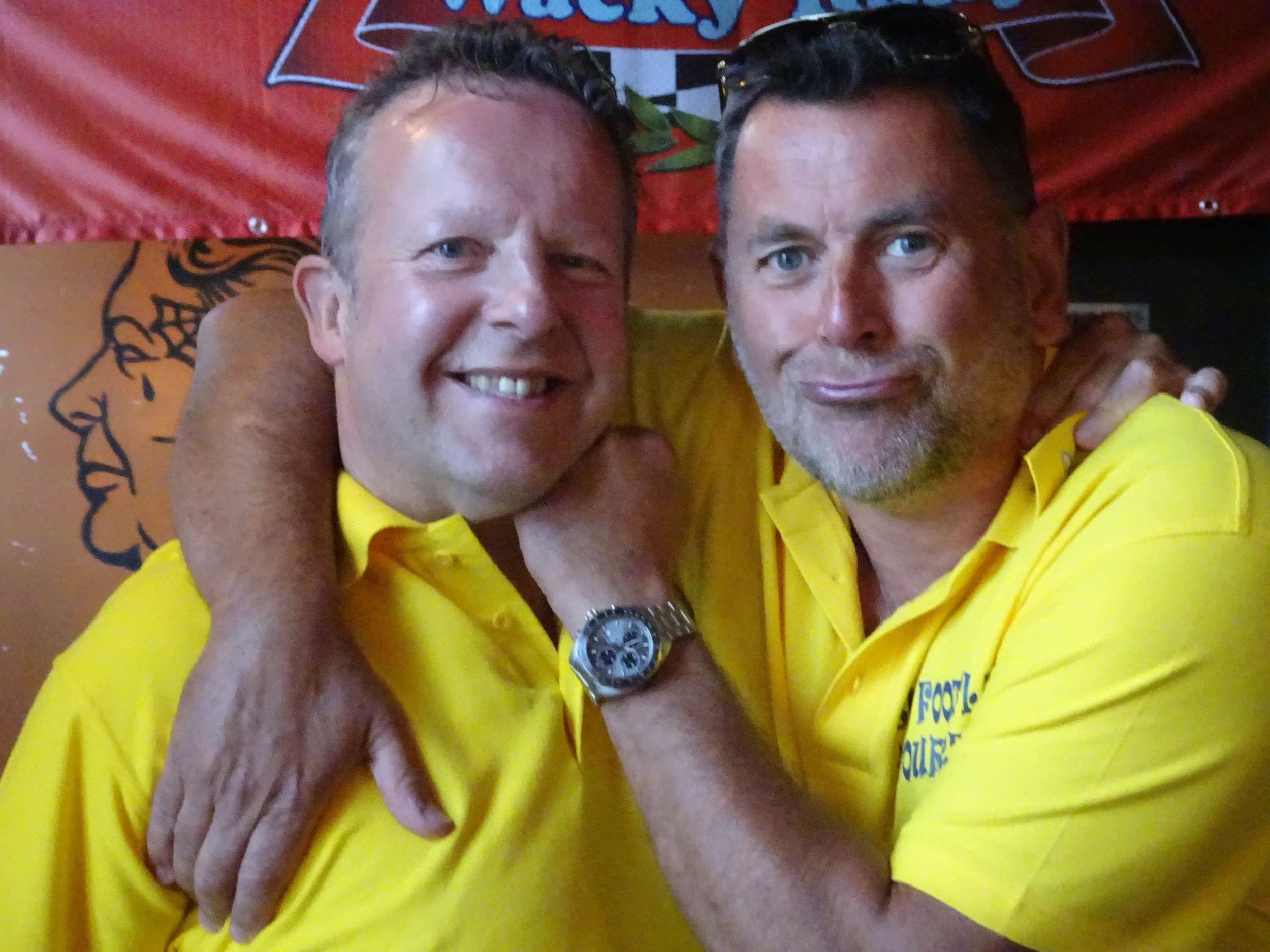 Two men wearing yellow shirts smiling and hugging each other, with a red background and a cartoon face drawing on the wall behind them.