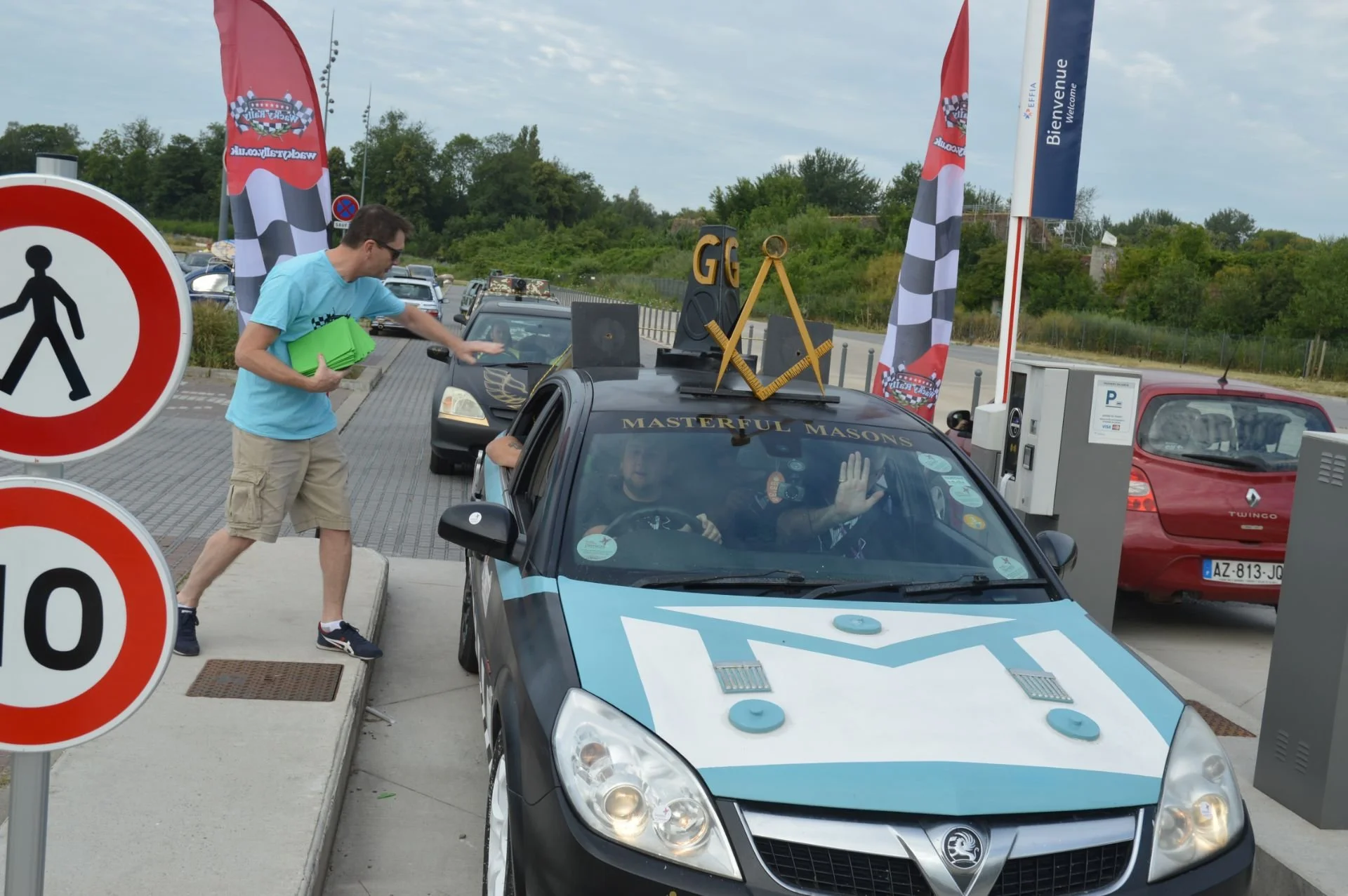 A man in a light blue shirt and khaki shorts is handing a green folder to a driver sitting in a police car with a blue and white police design. The police car has a sign on top that reads 'Masterful Masons' and is decorated with a builder's square, c
