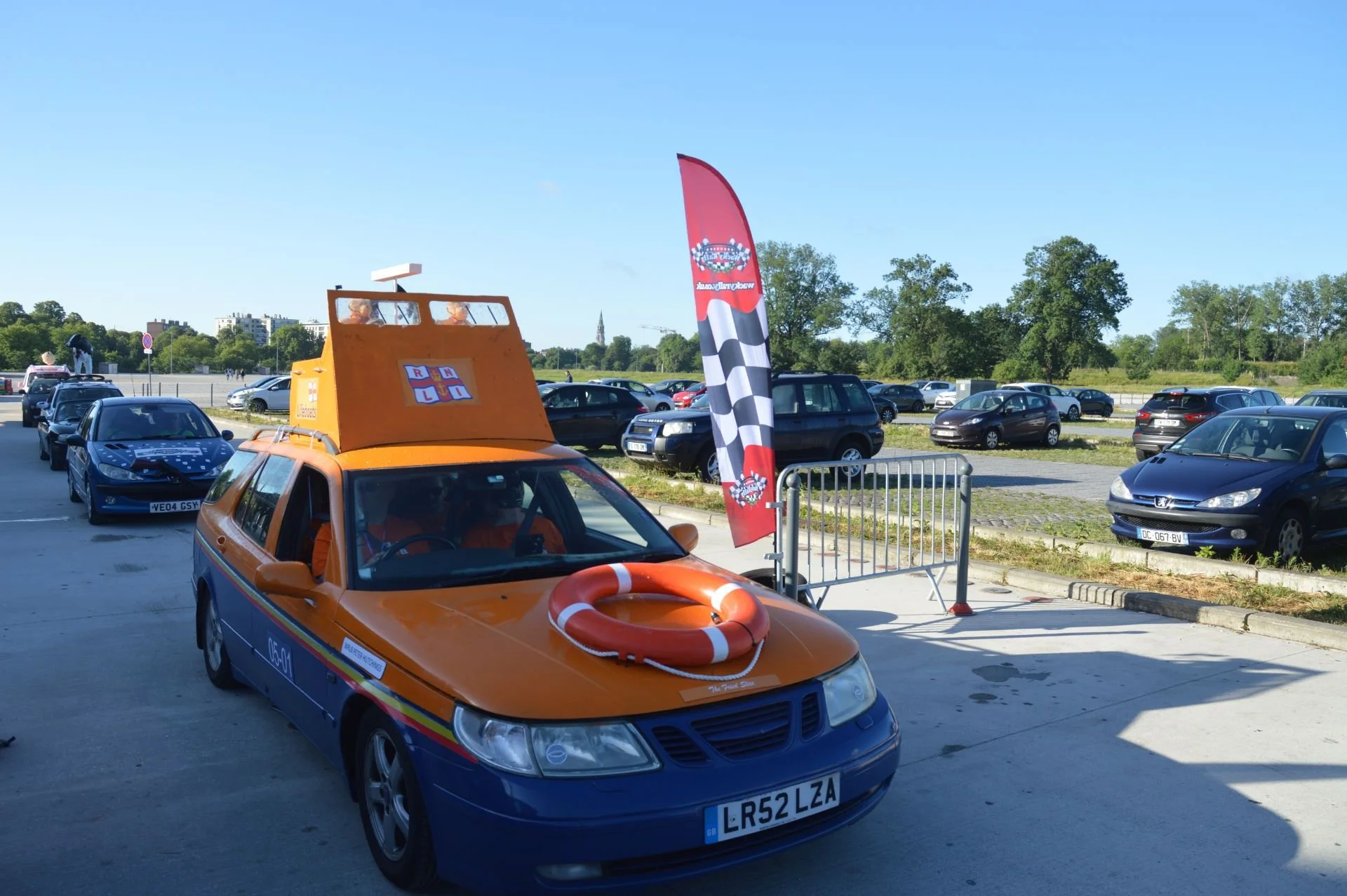 A car decorated as a lifeguard vehicle with a life preserver on the hood, parked in a lot next to a red and black checkered flag and a barrier, with other cars and greenery in the background.