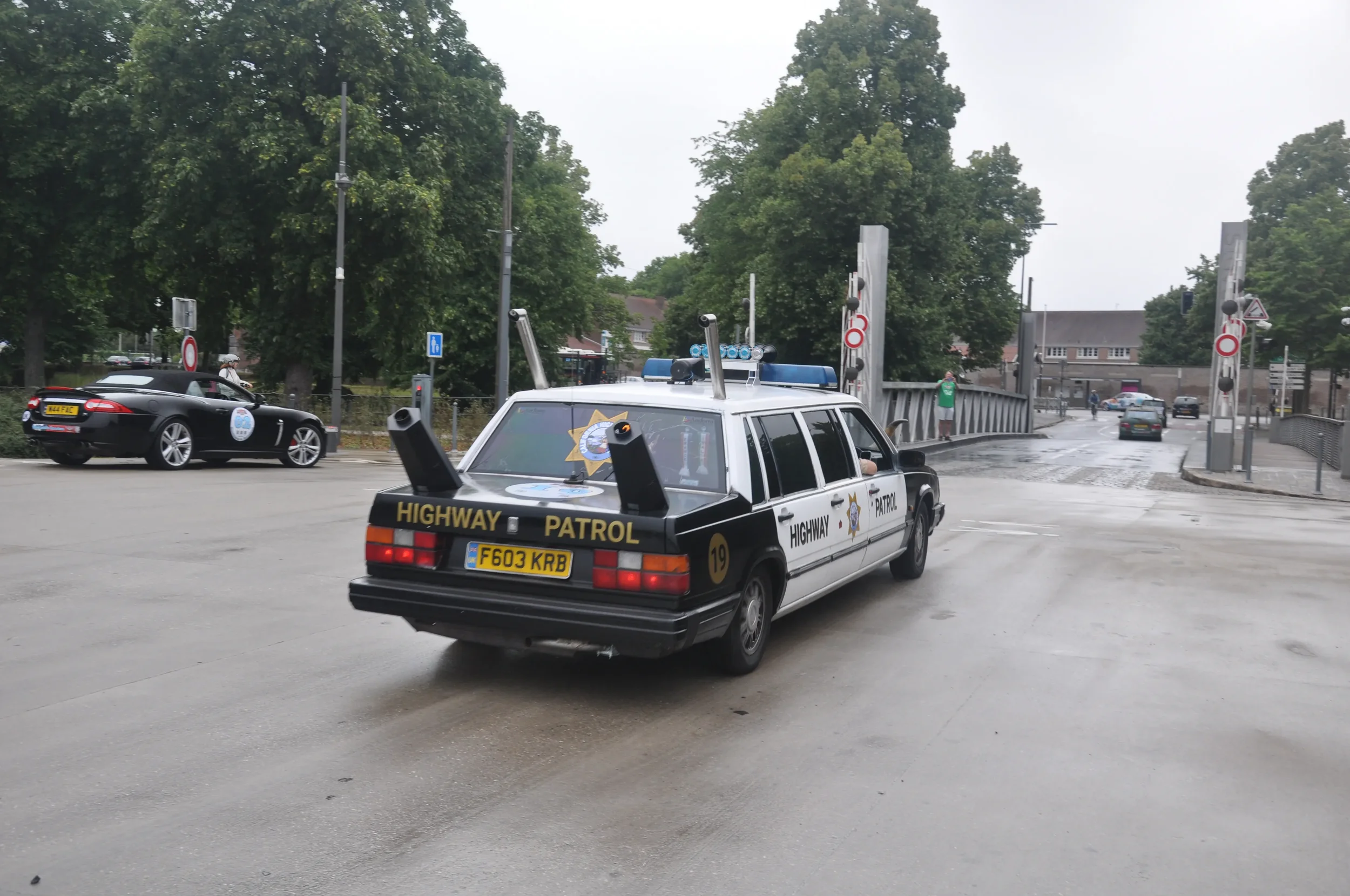 A highway patrol police car driving on a wet road near a bridge, with trees and a few cars in the background.