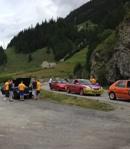 People gathered around several colorful cars parked on a gravel road by hills and rocky cliffs in a mountainous area.