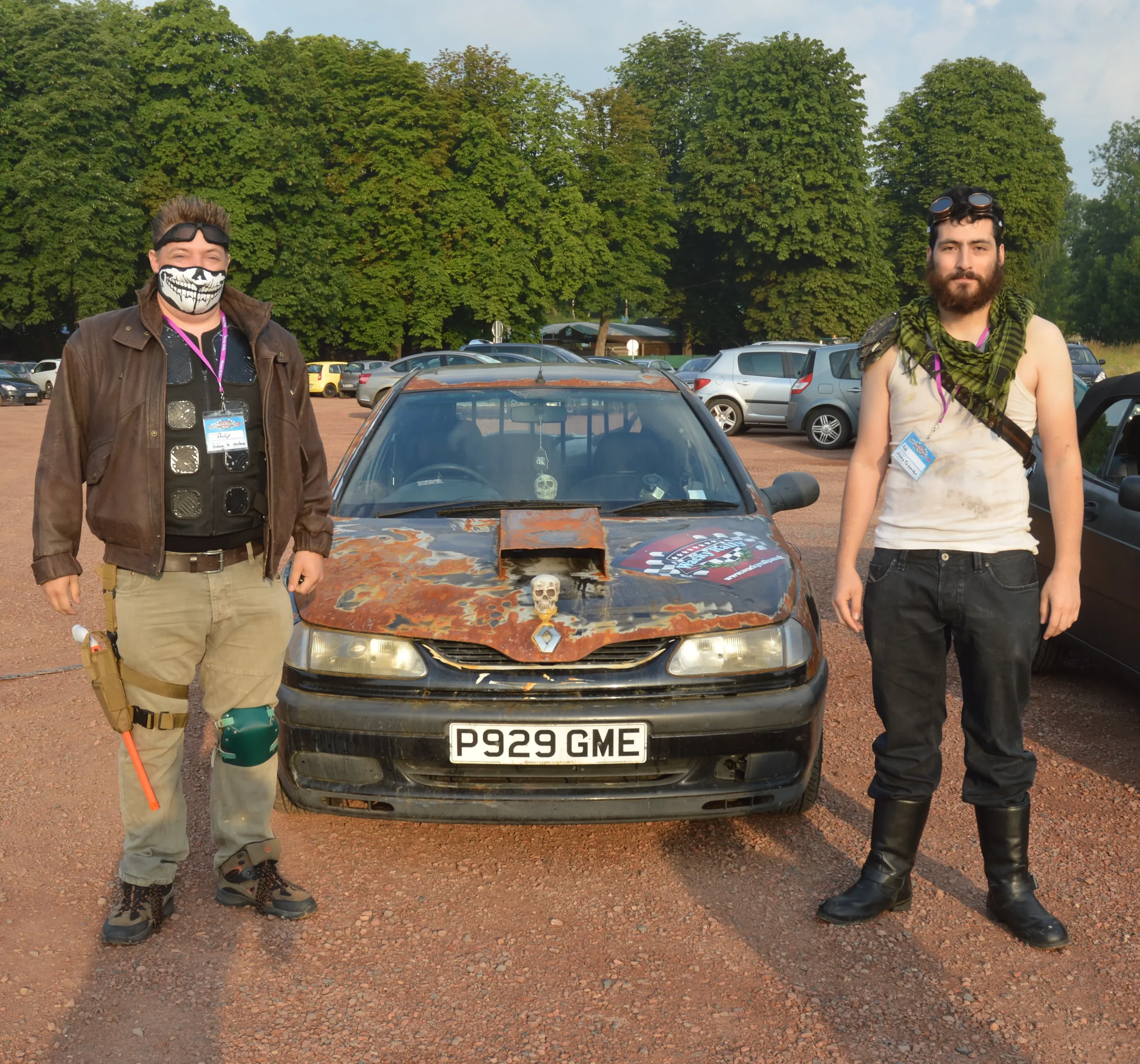 Two men dressed in cosplay standing beside a weathered, rusted car with a skull decoration on the hood, parked in a parking lot with trees in the background.