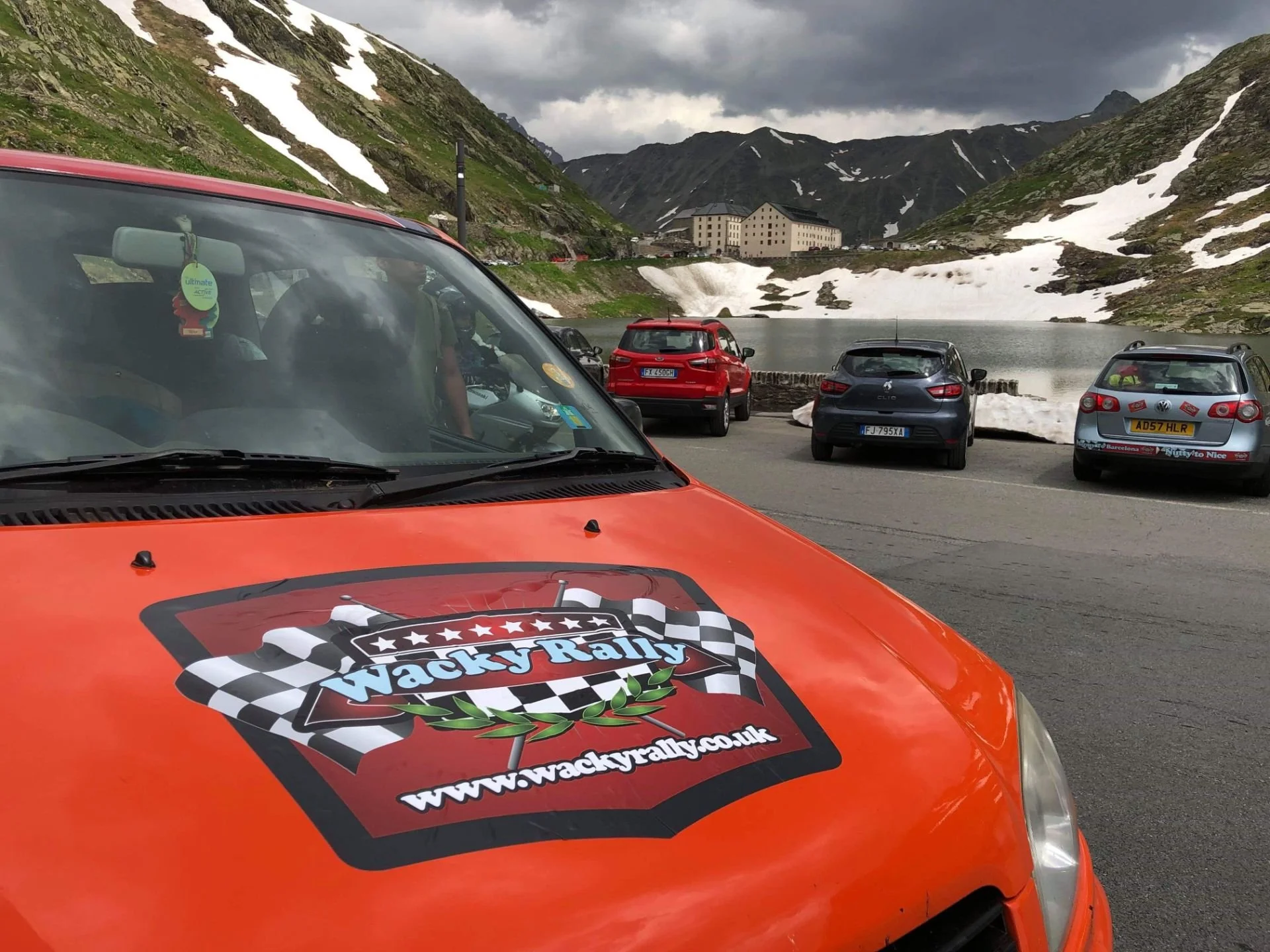 Orange rally car with WackyRally logo on the hood, parked near a mountain lake with snow patches, surrounded by other parked cars and mountain scenery.