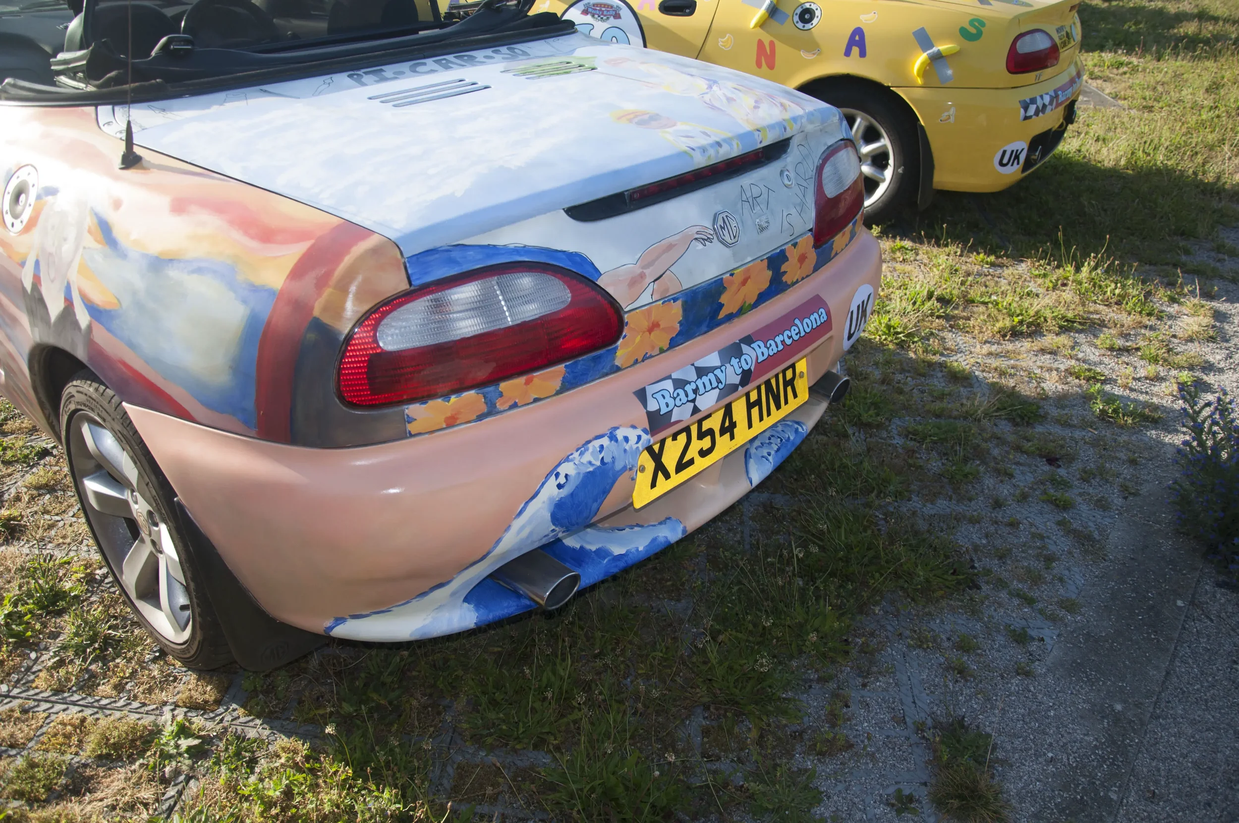 A painted car with artwork on the rear and sides, featuring a mermaid drawing and floral designs. The car has a sticker that says 'Darny to Barcelona' and a yellow UK license plate with the number X254 HNR. There are other decorated cars in the backg