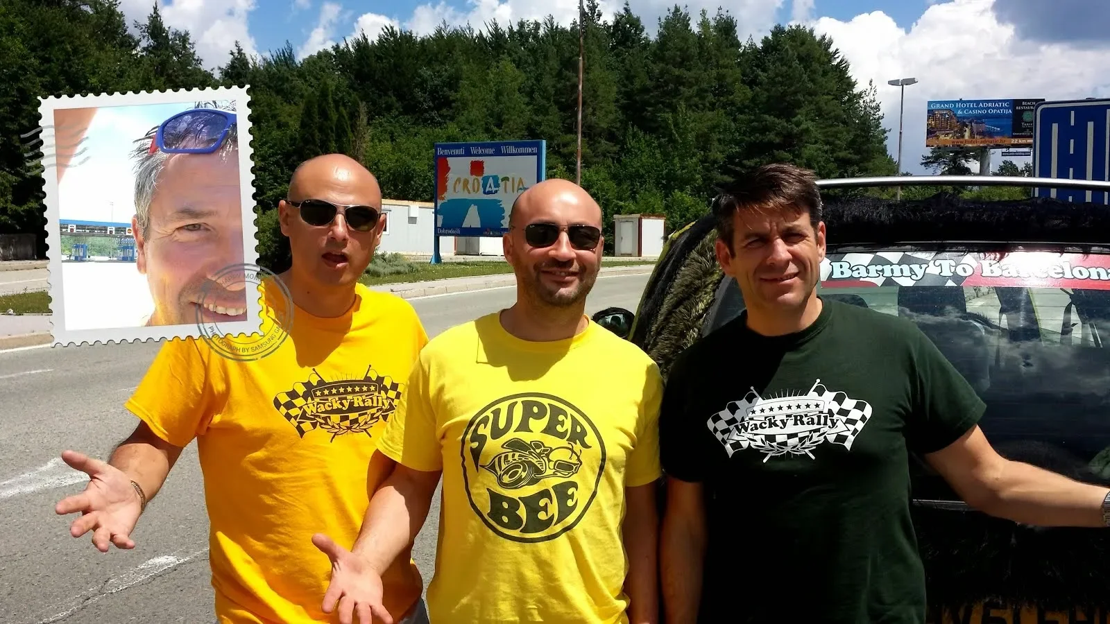 Three men standing on the side of a road, with a black vehicle behind them, in front of a sign that says "Croatia" and a billboard for a hotel and casino. The men are wearing T-shirts with rally-themed designs and sunglasses, smiling and gesturing to