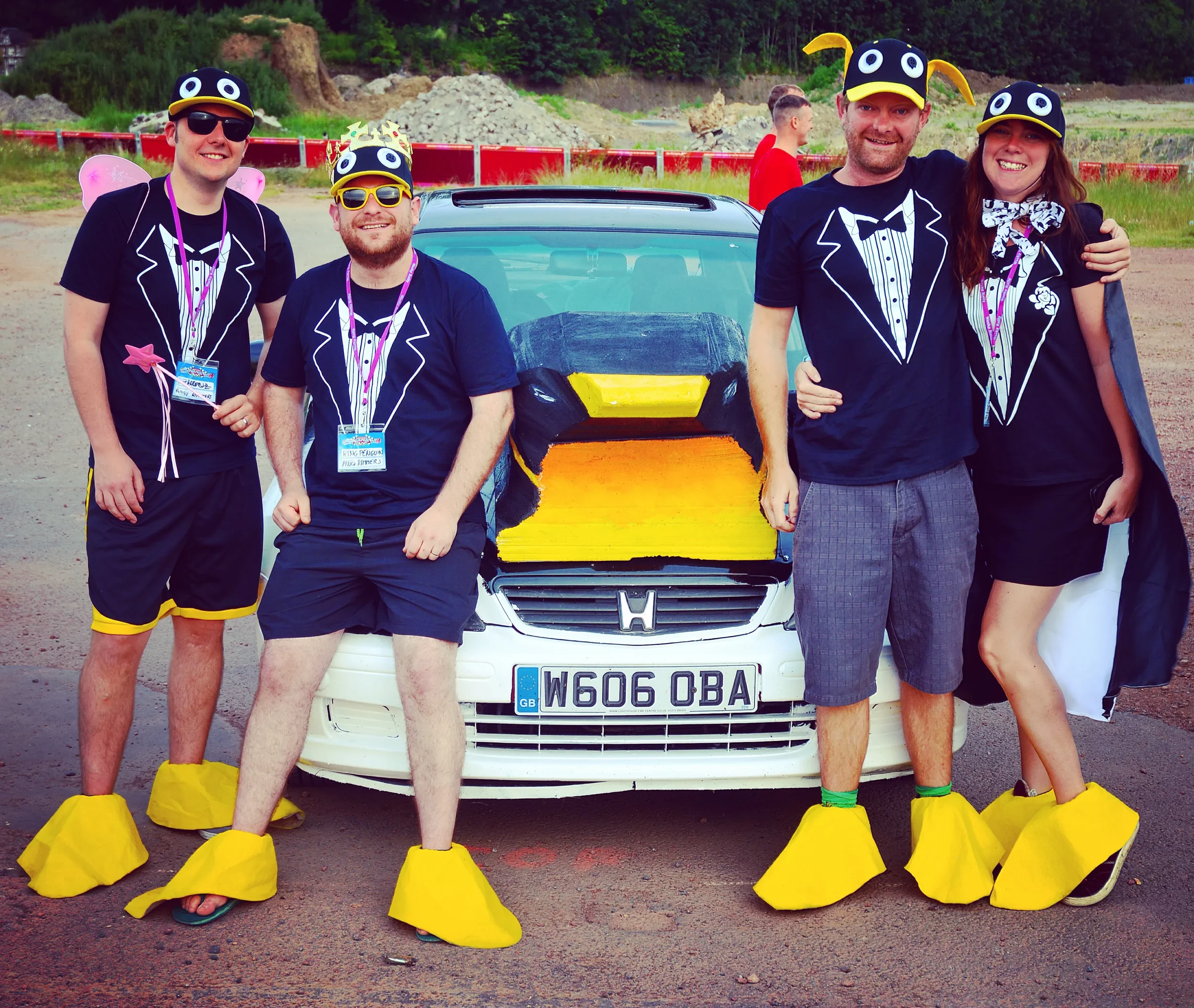 Group of five people dressed in whimsical costumes standing in front of a painted car with a dragon face on the hood. They are wearing matching tuxedo-print shirts, yellow foot covers, and hats resembling cartoon ducks, with some also having fairy wi