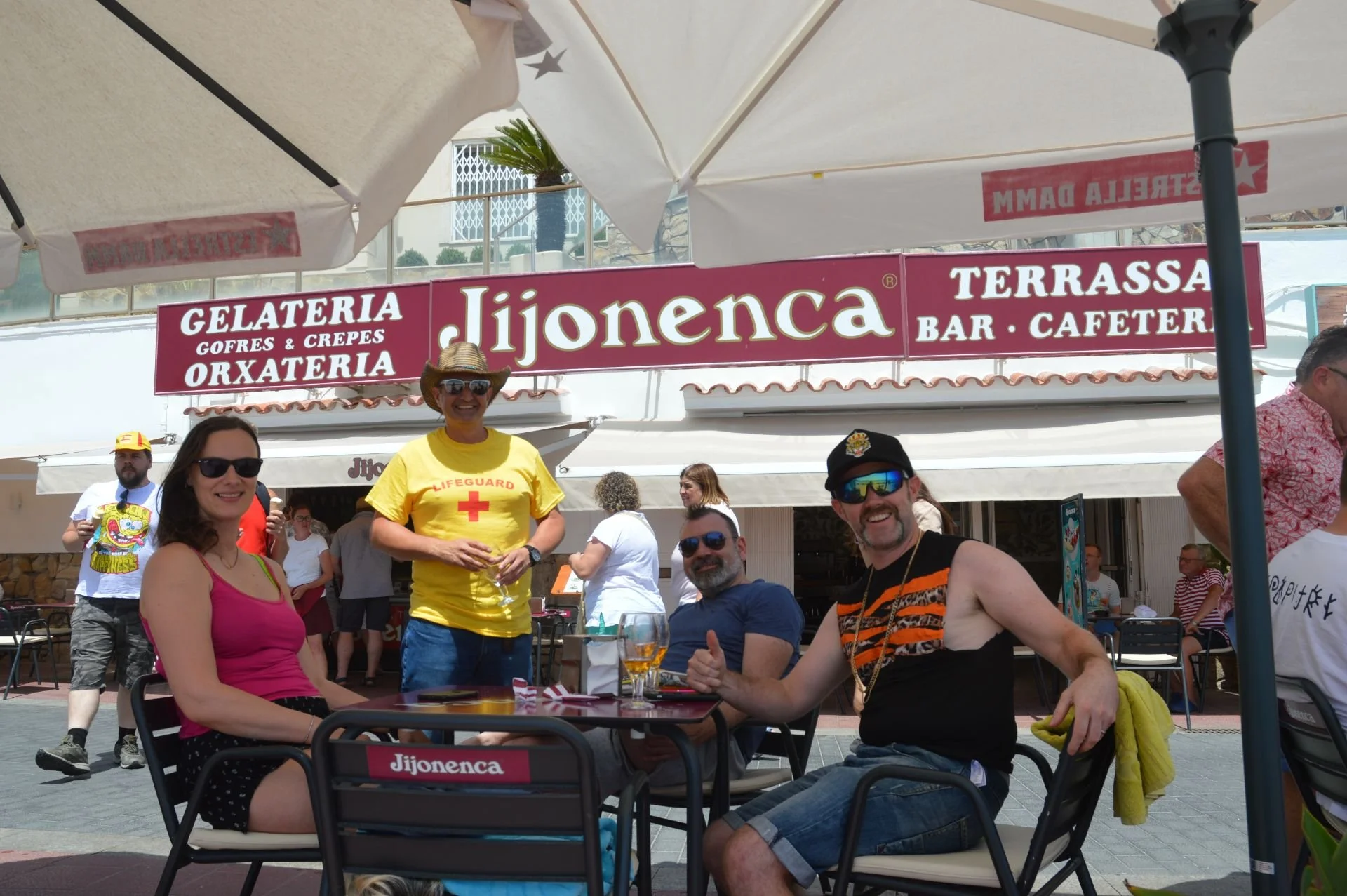 Group of friends sitting at a table outside a restaurant named Jijonenca, under umbrellas, with a muscle man, a woman in a pink top, and a man in a tank top giving thumbs up, with other people dining in the background.