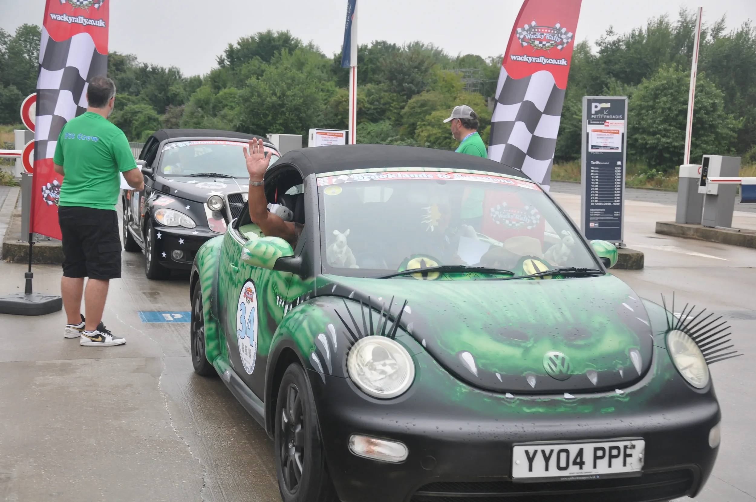 A green cartoon dragon painted Volkswagen Beetle with added eyelashes and dragon teeth on the front, parked at a rally event with people and other cars around.
