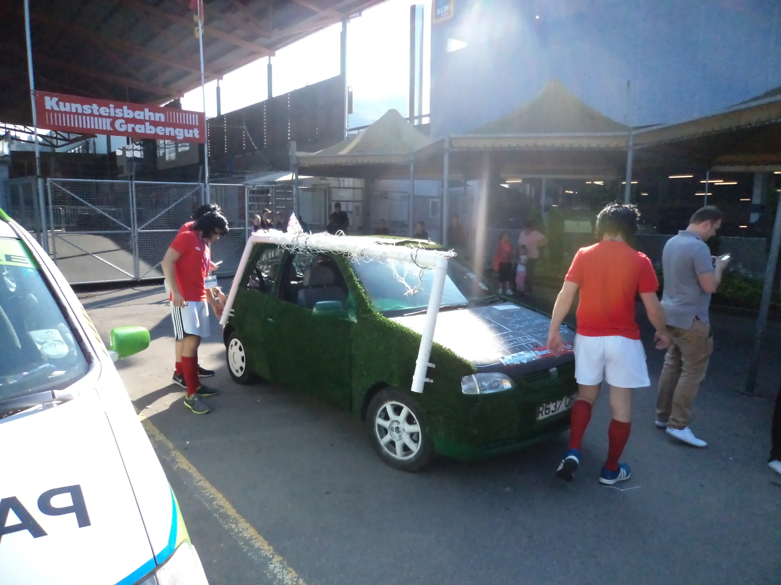 A small car covered in green artificial grass parked in a parking lot with four young men standing nearby. The car has a makeshift white goalpost on top and is situated under summer sunlight at an outdoor event.
