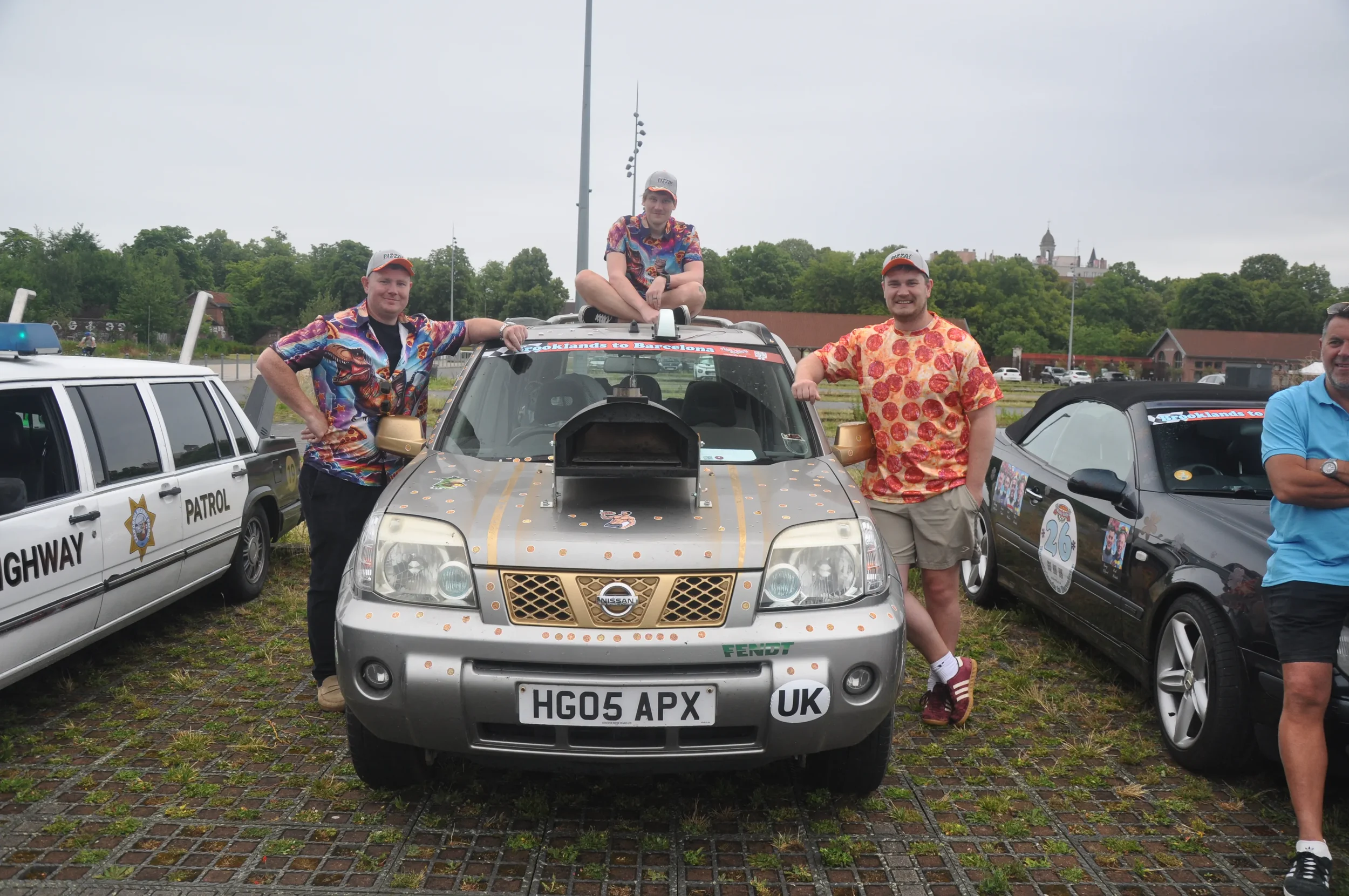 Group of men standing around a decorated silver Nissan SUV and two other cars, one resembling a police patrol car, at an outdoor event.