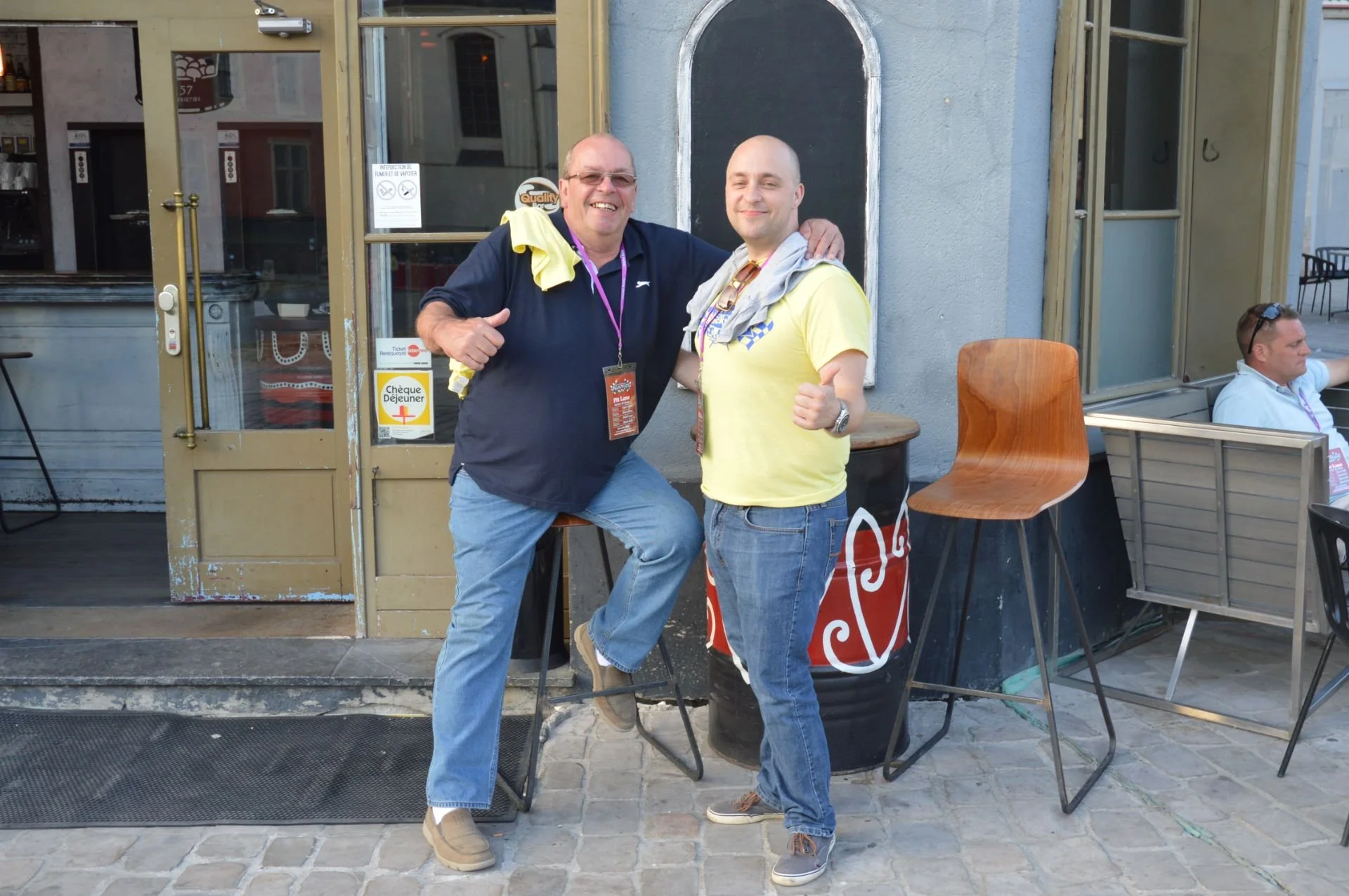Two men smiling and posing with thumbs up outside a restaurant, standing on a cobblestone sidewalk.