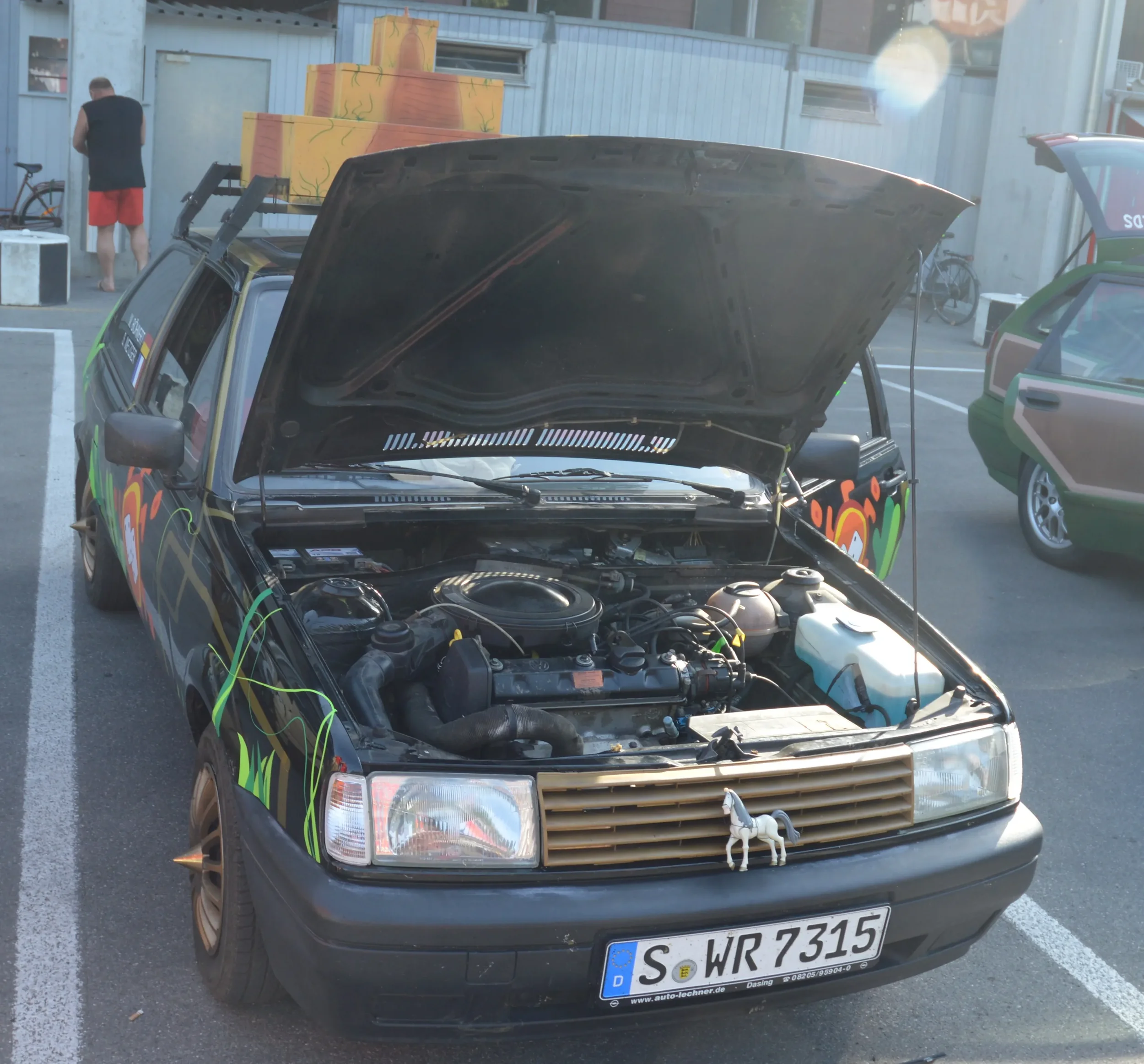 A black car with artwork on the sides has its hood open, revealing the engine. The front of the car displays a small horse figurine hanging from the grille. It is parked in a lot with other vehicles and a person in red shorts and a black shirt in the