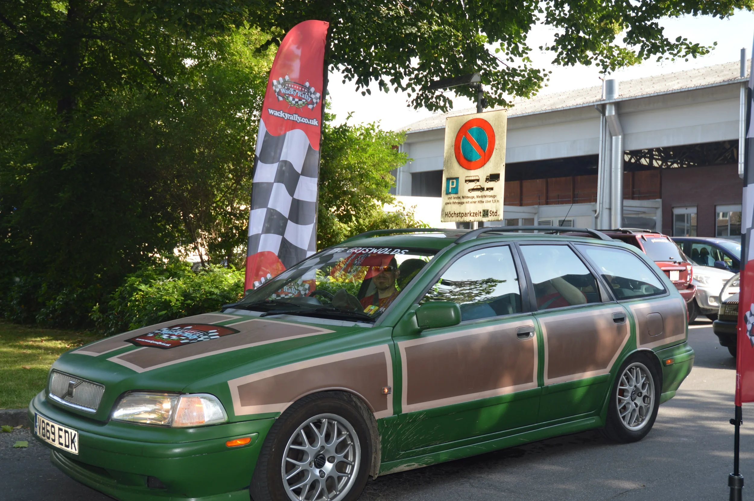 A green car with custom beige and green paintwork resembling a race car, parked outdoors near a tree and a parking lot with several other cars. Tall red and black checkered flags with the logo 'Wacky Rally' are on either side of the car.