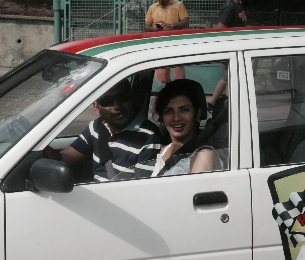 Two people sitting inside a white car, smiling at the camera, with a woman wearing a white top and a man in a striped shirt and sunglasses.