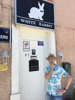 Person standing in front of a white door with a White Rabbit sign above it, wearing sunglasses and a tropical shirt.