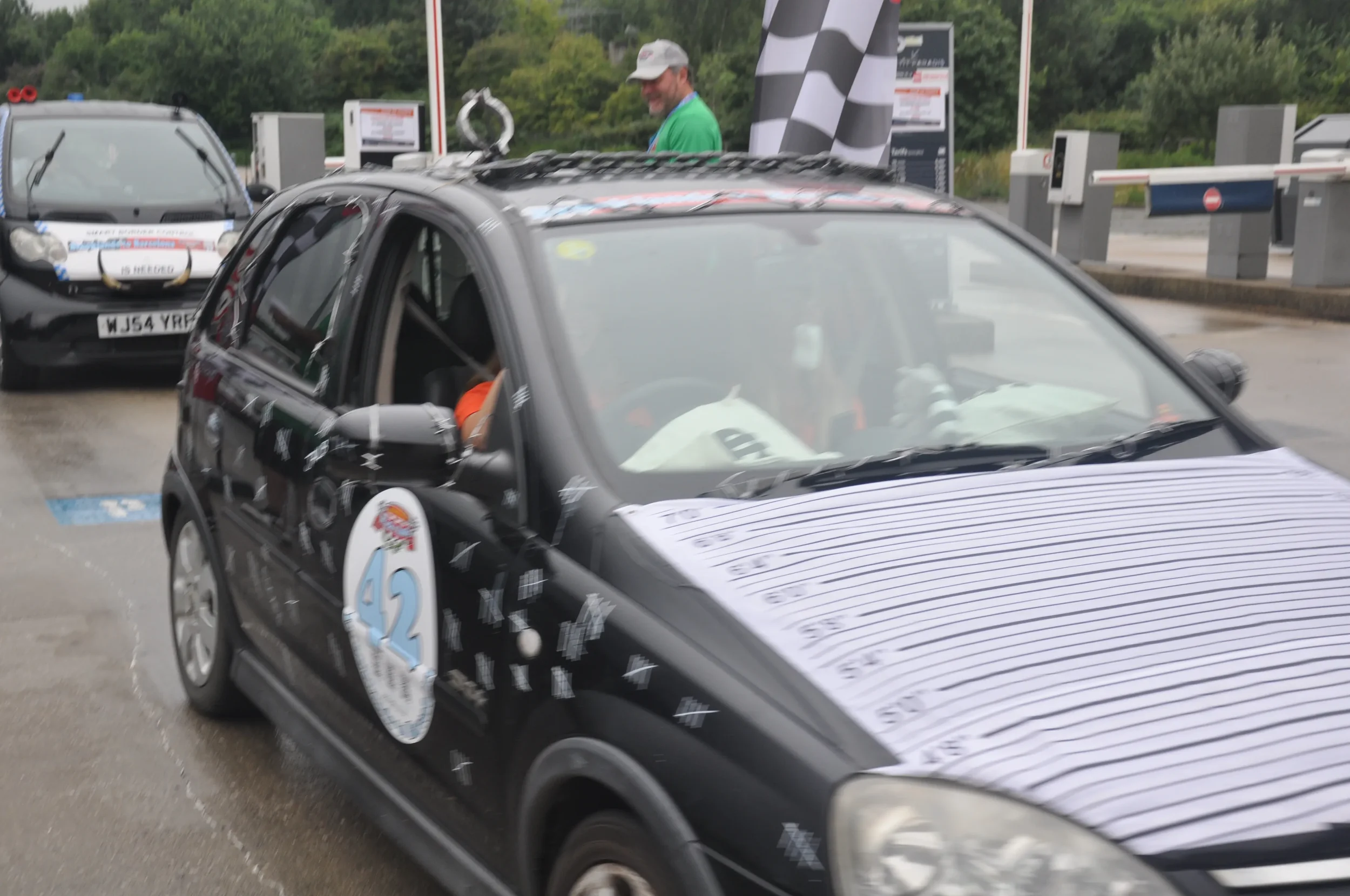 A black rally car with race markings and sponsor decals, parked at a race track finish line with a checkered flag and timing sheet on the hood. A man is smiling in the background near other cars.