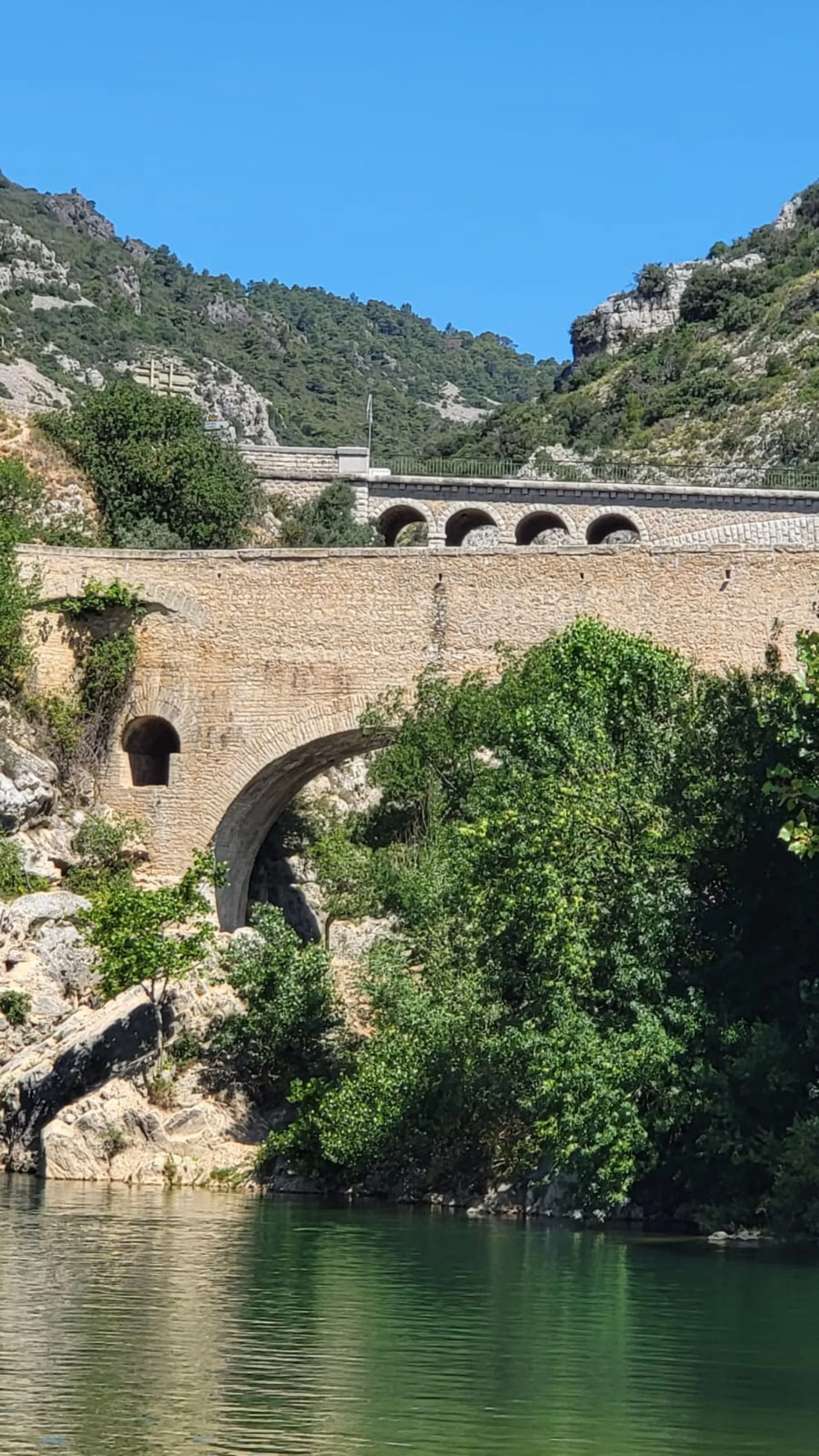 Scenic view of a stone bridge with multiple arches over a river, surrounded by green trees and mountains under a clear blue sky.