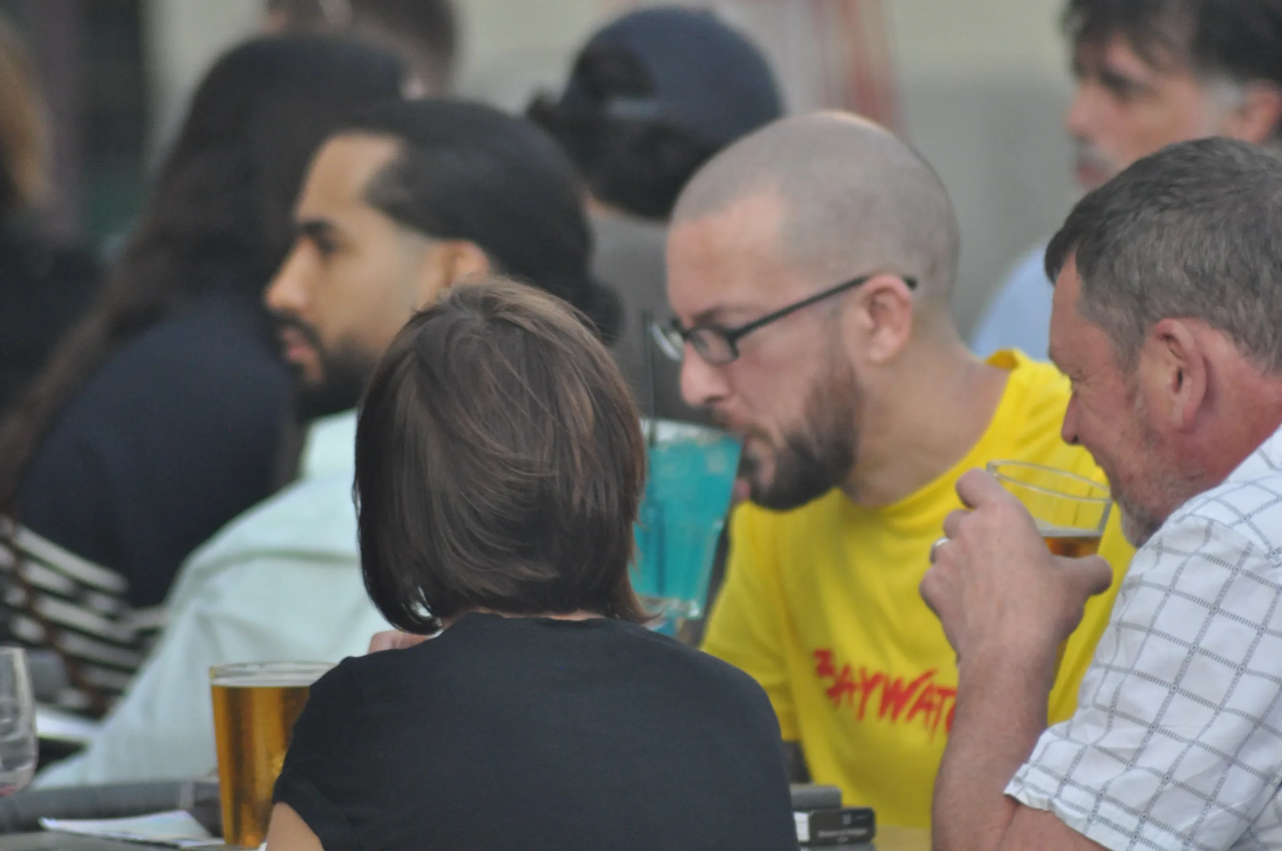 Multiple people seated at a long table, some drinking beers, engaging in a social gathering or event.