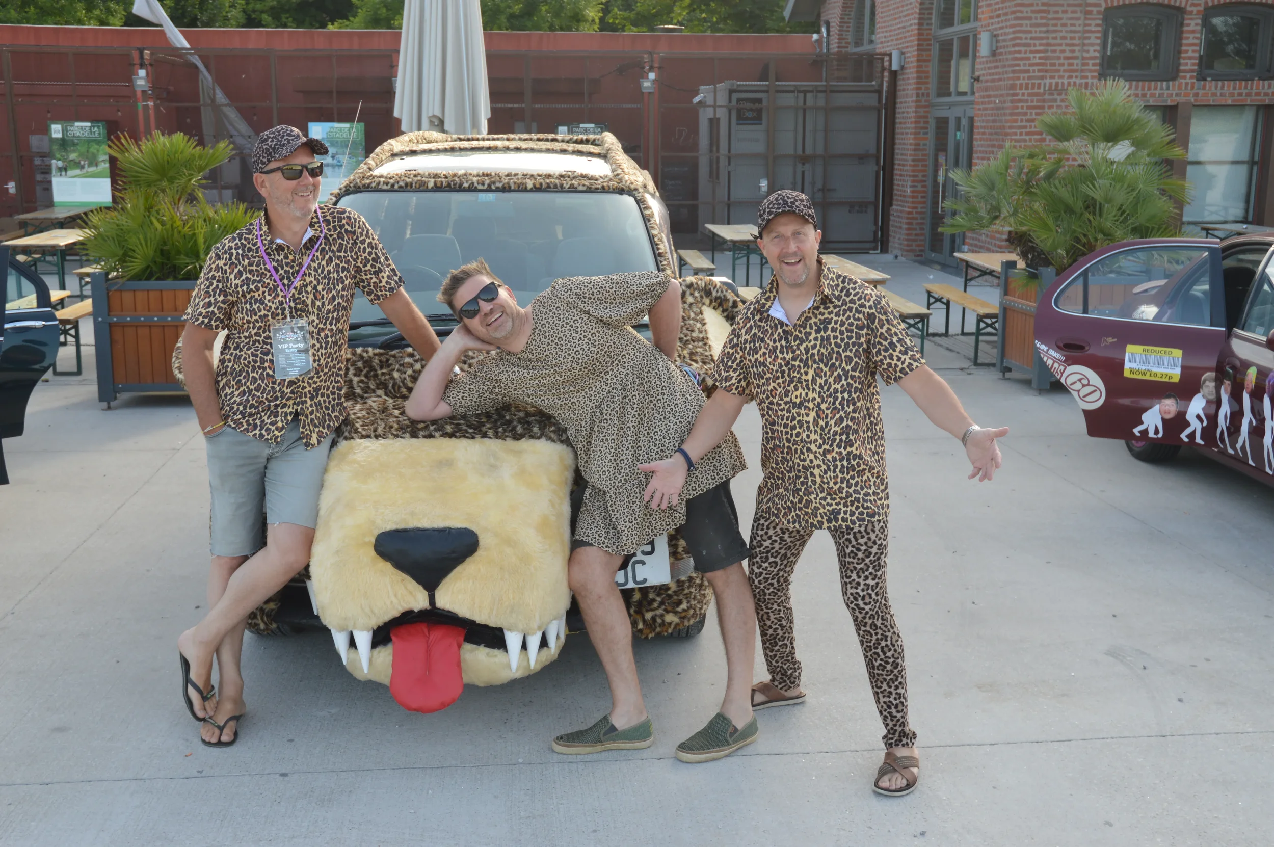 Four men wearing leopard print outfits posing around a car to resemble a big cat at an outdoor event.