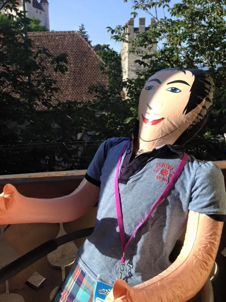A humanoid balloon figure with a paper mask face, wearing a gray polo shirt with a pink lanyard, sitting on a balcony with trees and rooftops in the background.