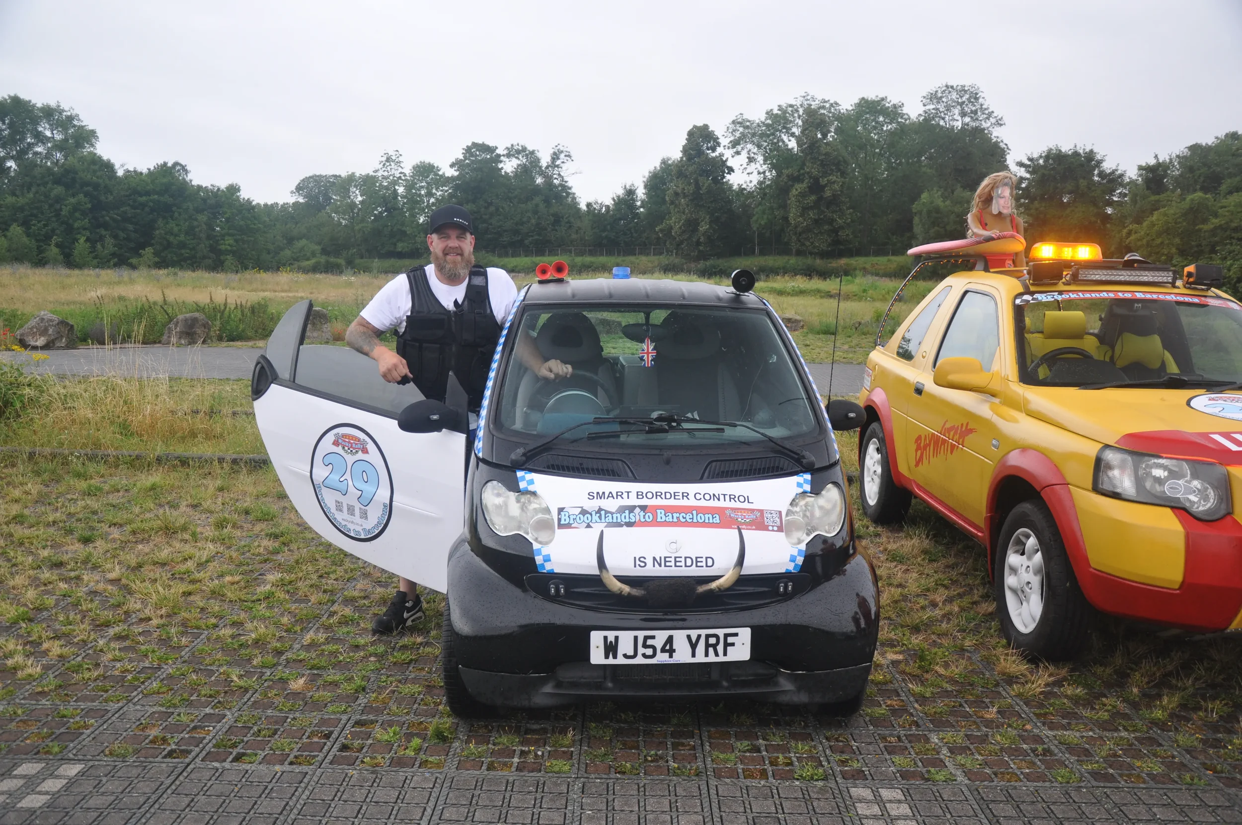 A man stands next to a small black electric car with a sign that says 'Smart Border Control' and 'Brooklands to Barcelona,' parked on a paved area with grassy fields and trees in the background. The car has a license plate 'WJ54 YRF' and a person ins