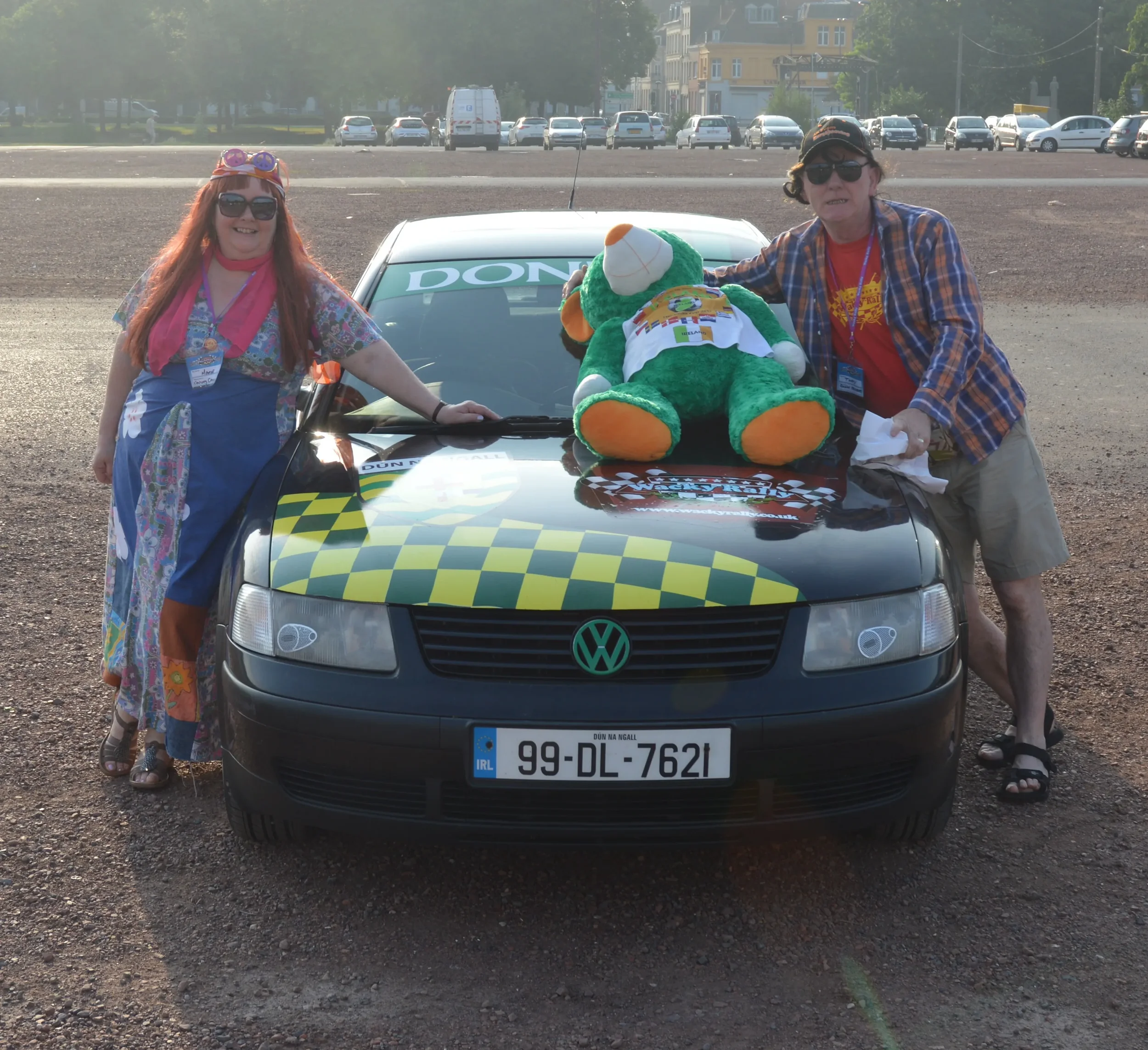 Two people standing next to a black Volkswagen car decorated with checkered yellow and black pattern, with a large green teddy bear dressed in a colorful outfit on the hood. The woman on the left is wearing sunglasses, a colorful dress with a pink sc