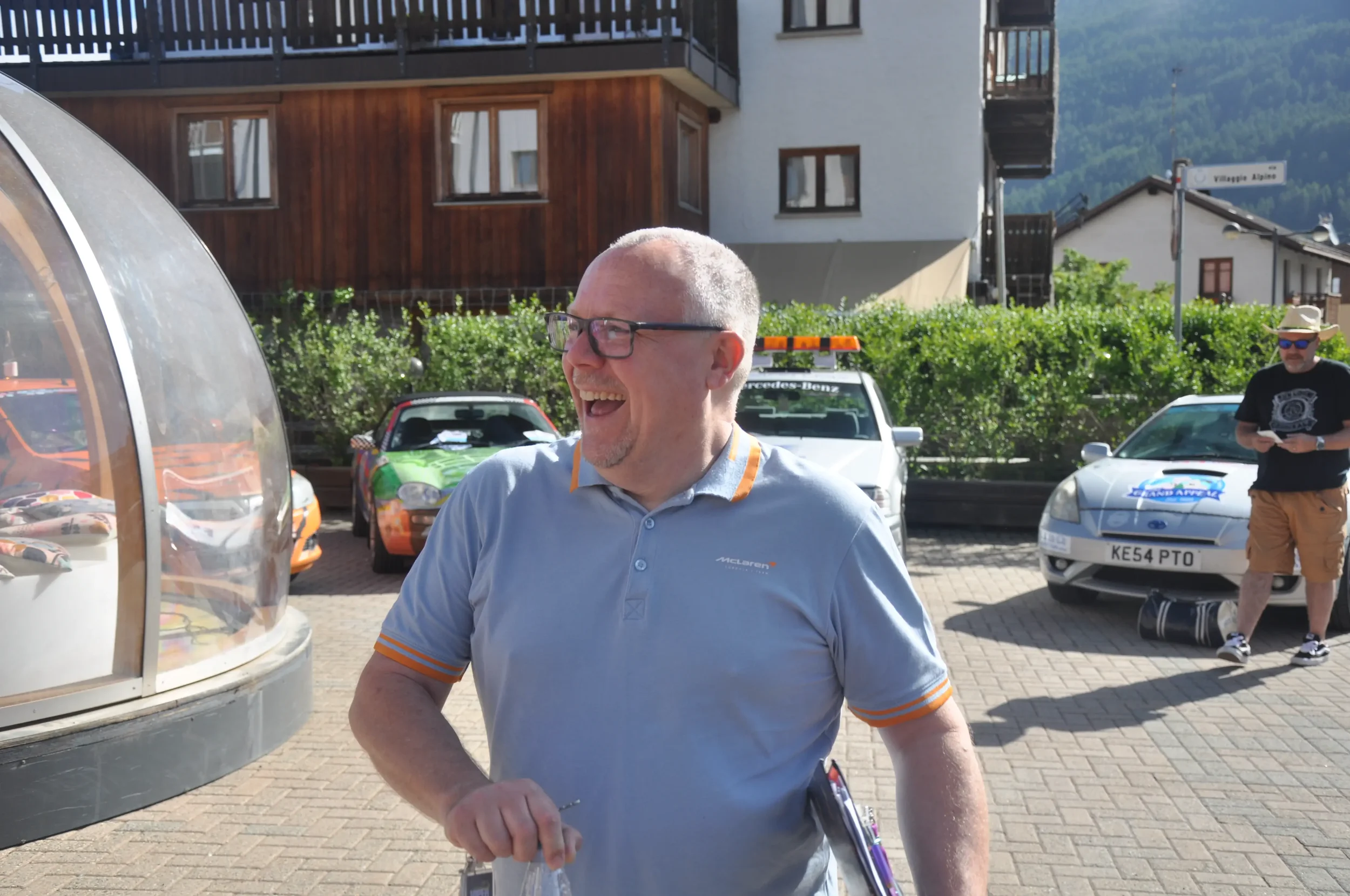 A man with glasses and a gray polo shirt smiling and laughing outdoors in a parking lot with colorful cars and another man in the background using a phone, surrounded by buildings and greenery.