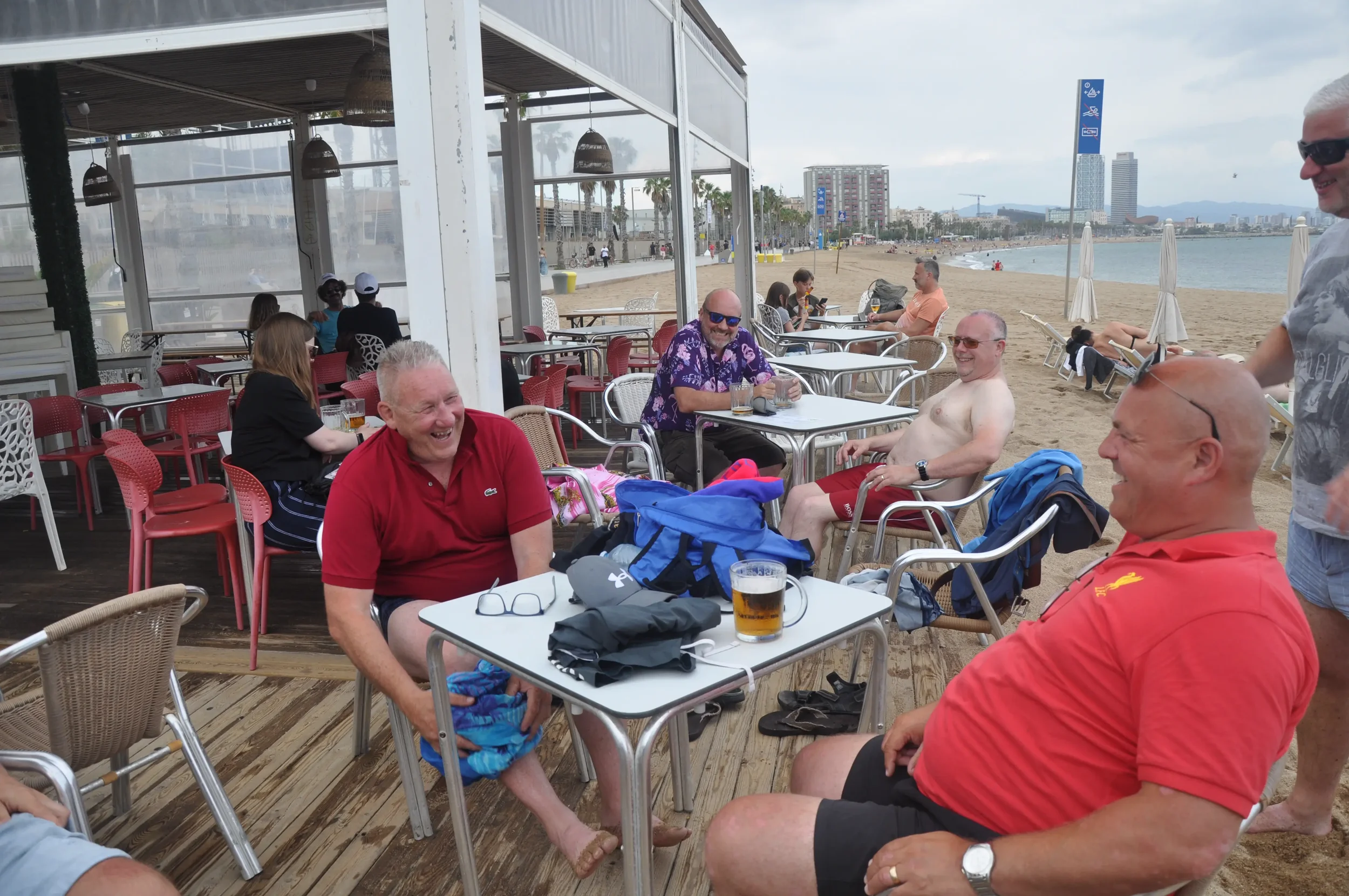 Group of people enjoying drinks and conversation at an outdoor beachside restaurant, with sandy beach and ocean in the background.
