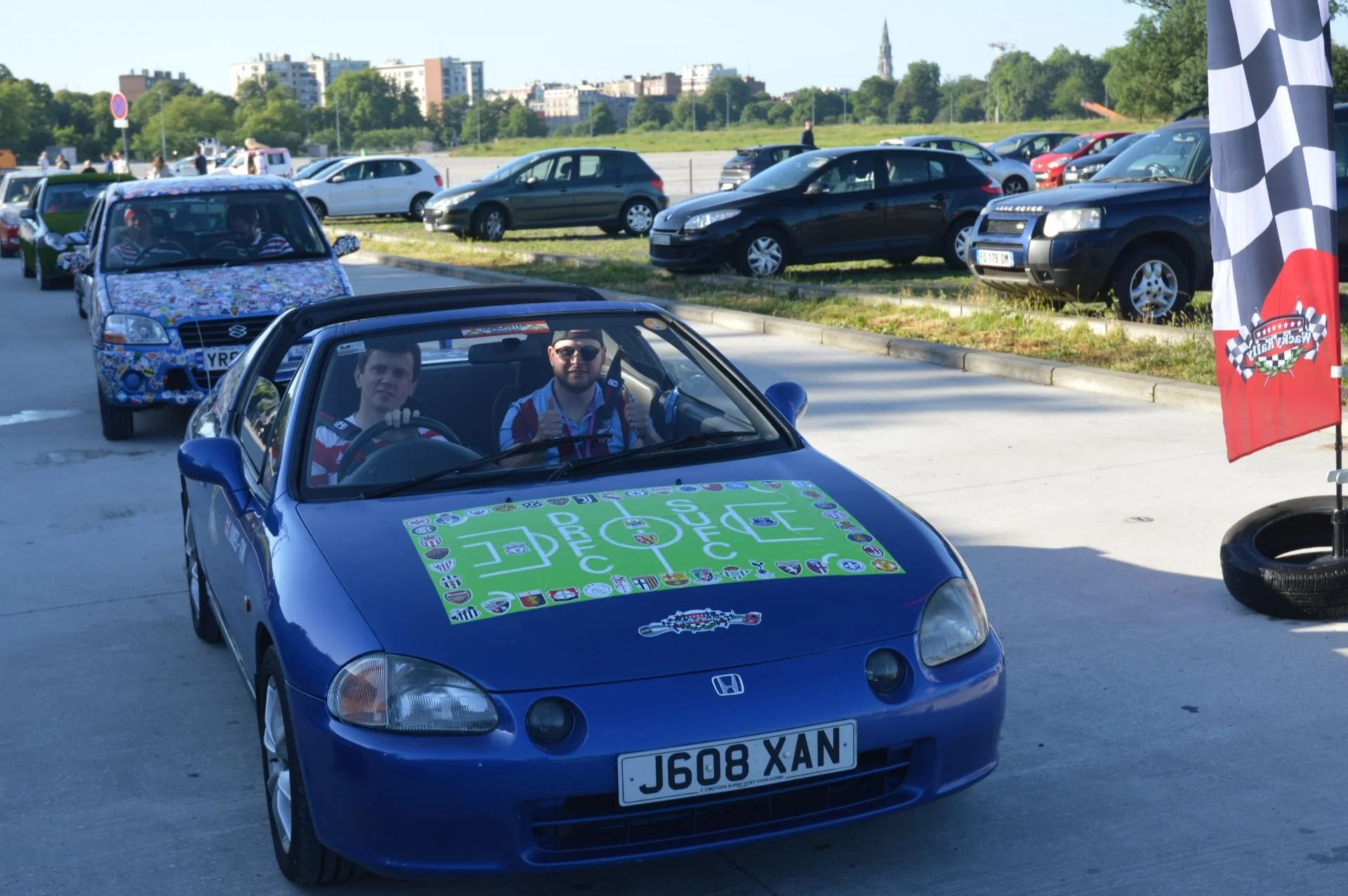 A blue Honda sports car with a race-themed decal on the hood, parked at a car event, with two men inside giving thumbs up. The background shows other parked cars and a cityscape with trees and buildings.