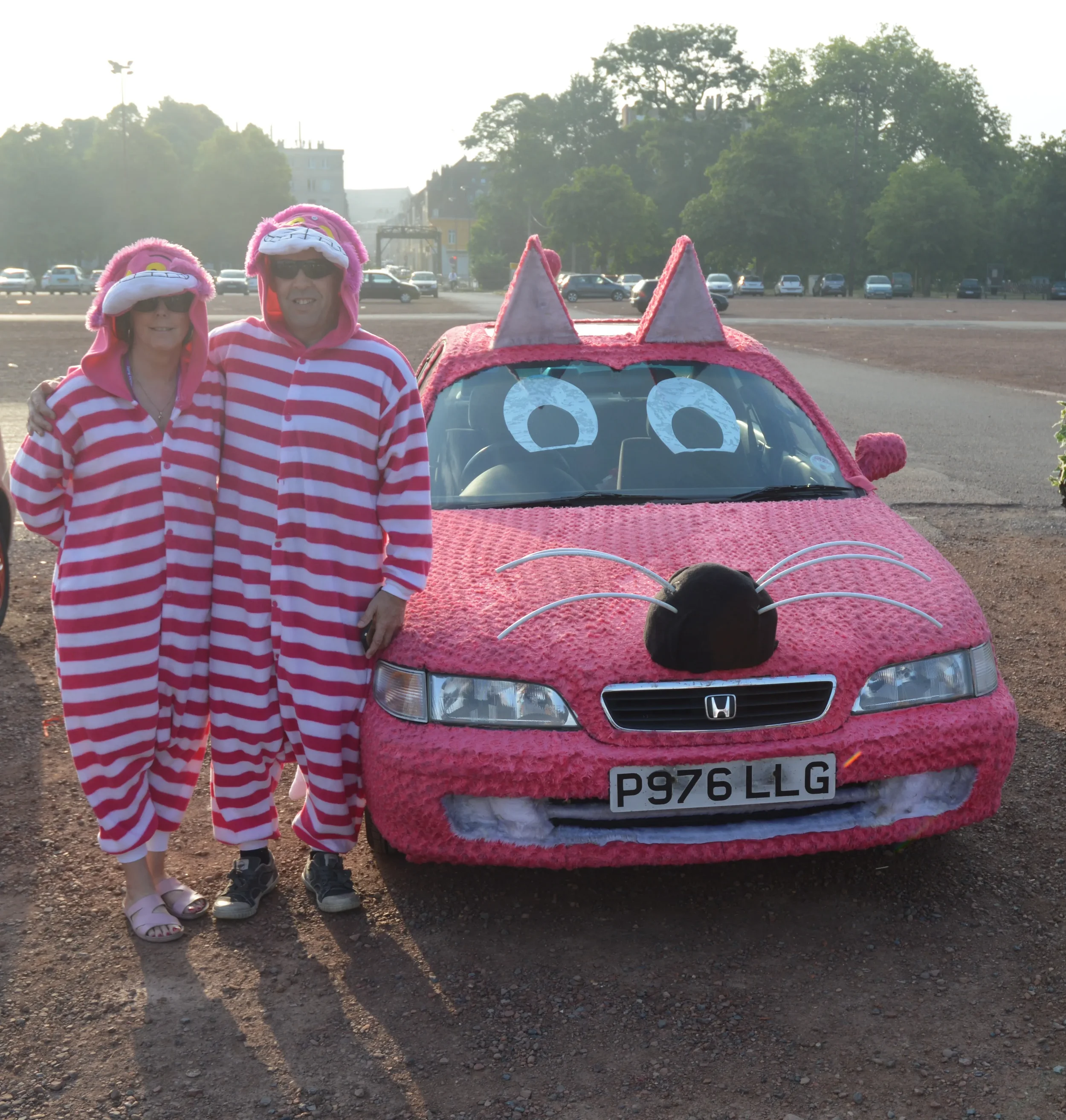 A man and woman in pink and white striped pajamas stand next to a car decorated as a pink cat with eyes, a nose, whiskers, and ears, at dusk in an open parking lot.
