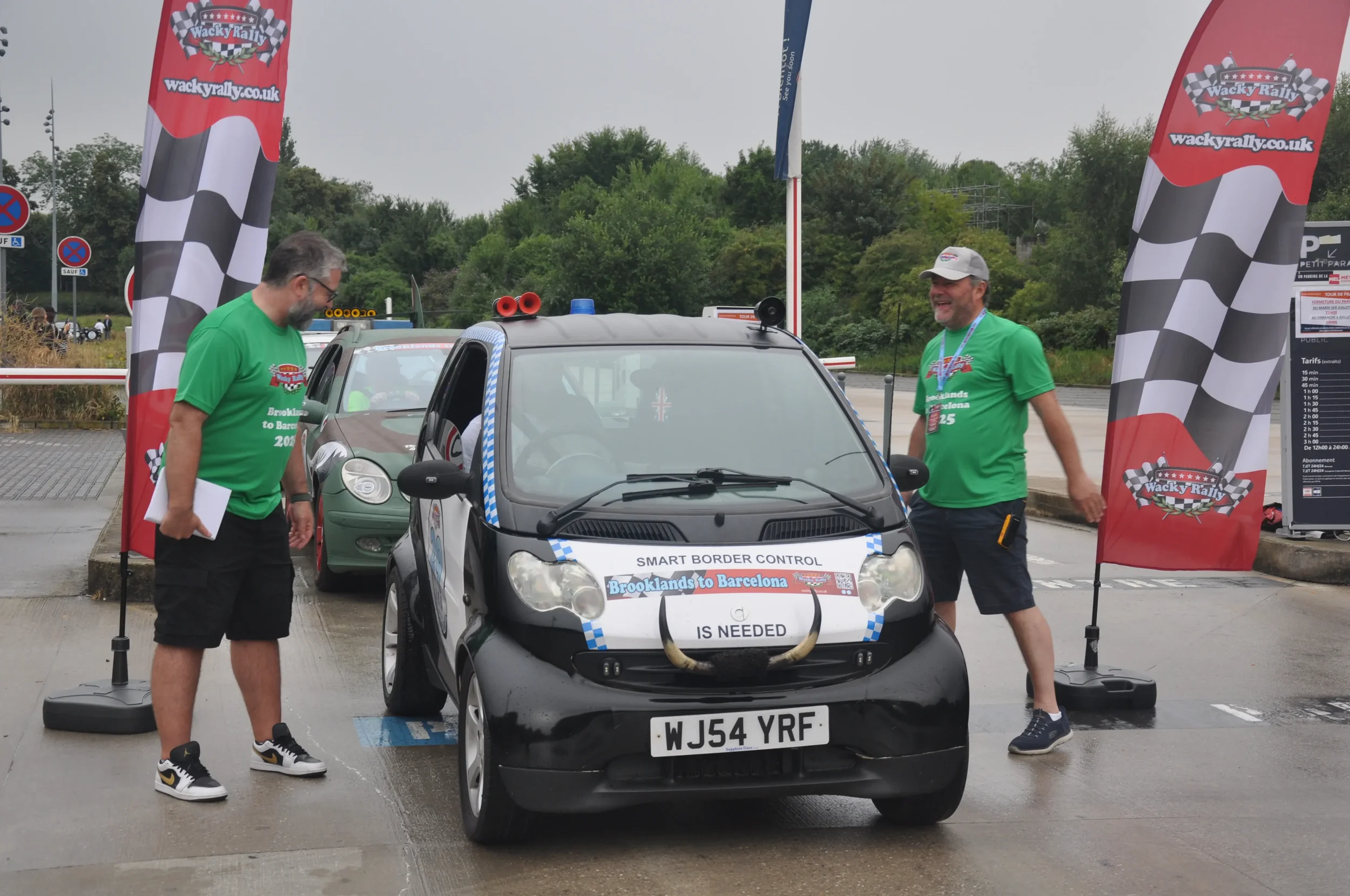 Two men in green shirts standing next to a small black car with a sign that reads 'Smart Border Control, Brooklands to Barcelona is needed', decorated with a bulls' horns attached to the front bumper. They are at a wacky rally event with red and blac