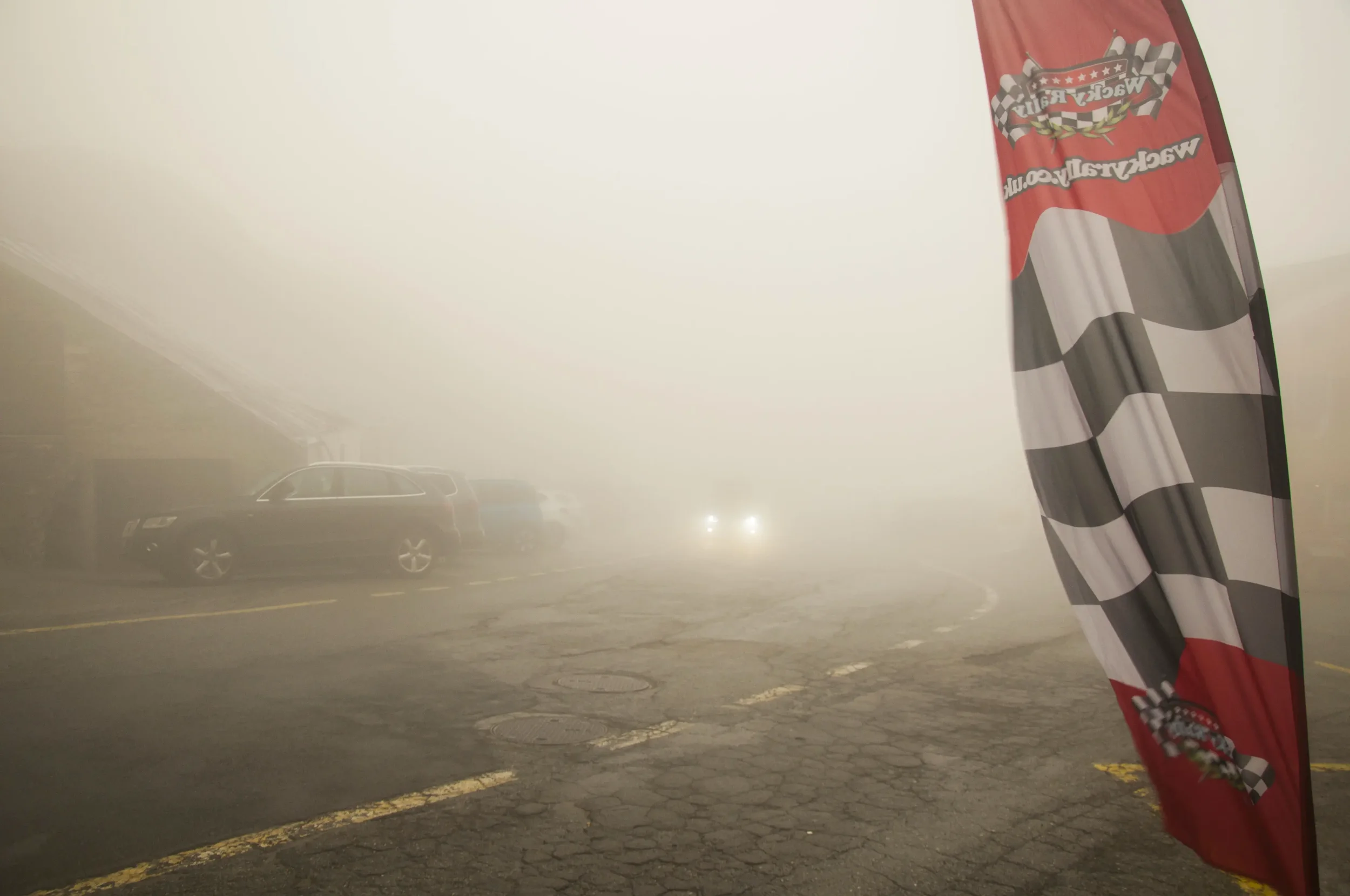 Foggy parking lot with a large checkered flag on a pole and a dark station wagon in the background, with headlights barely visible through the fog.