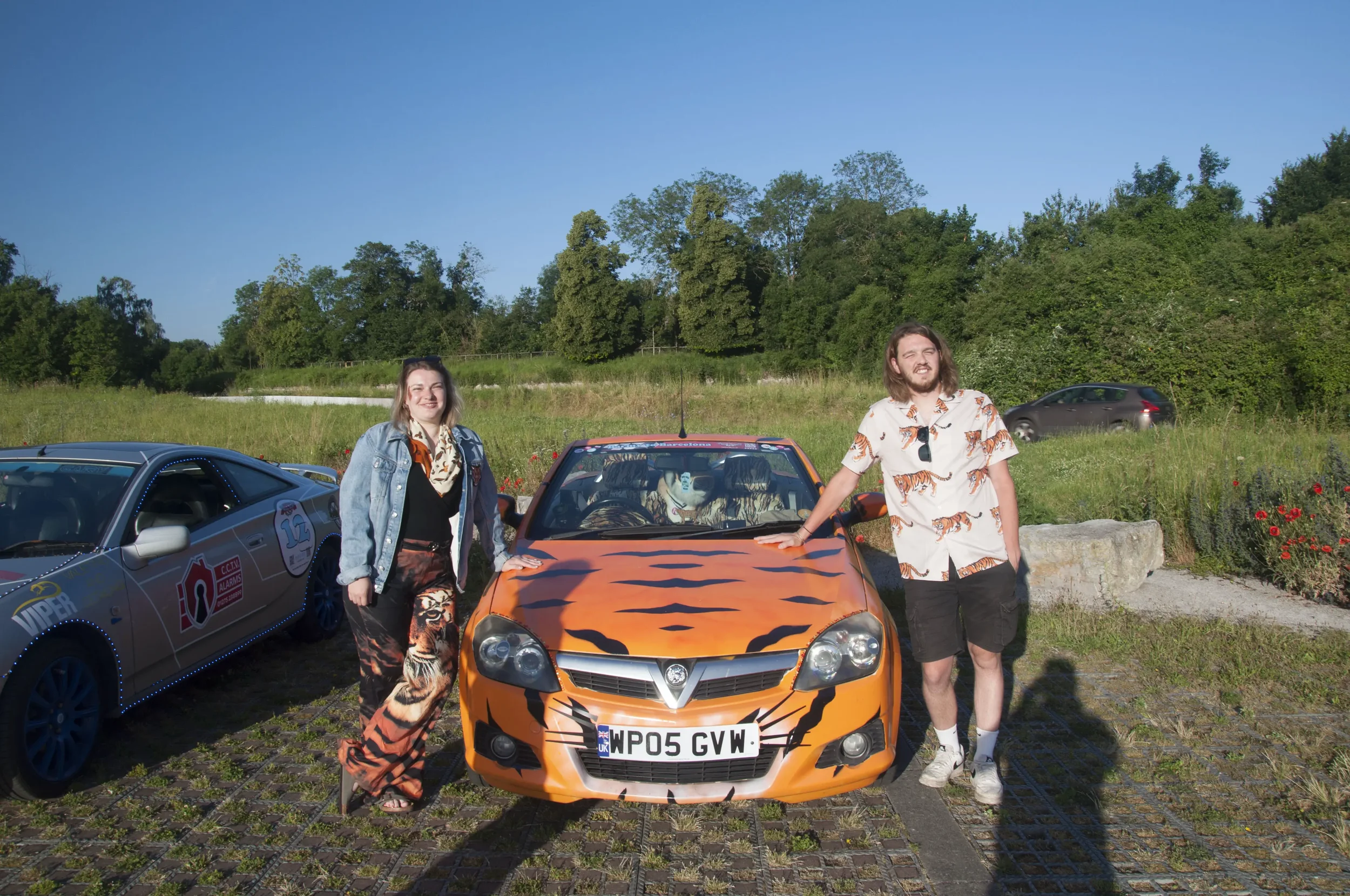 A young woman and a young man are standing beside a small orange car with a tiger face paint design, in a parking lot with grass and trees in the background. The woman is smiling, wearing tiger-patterned pants and a denim jacket, while the man is lea