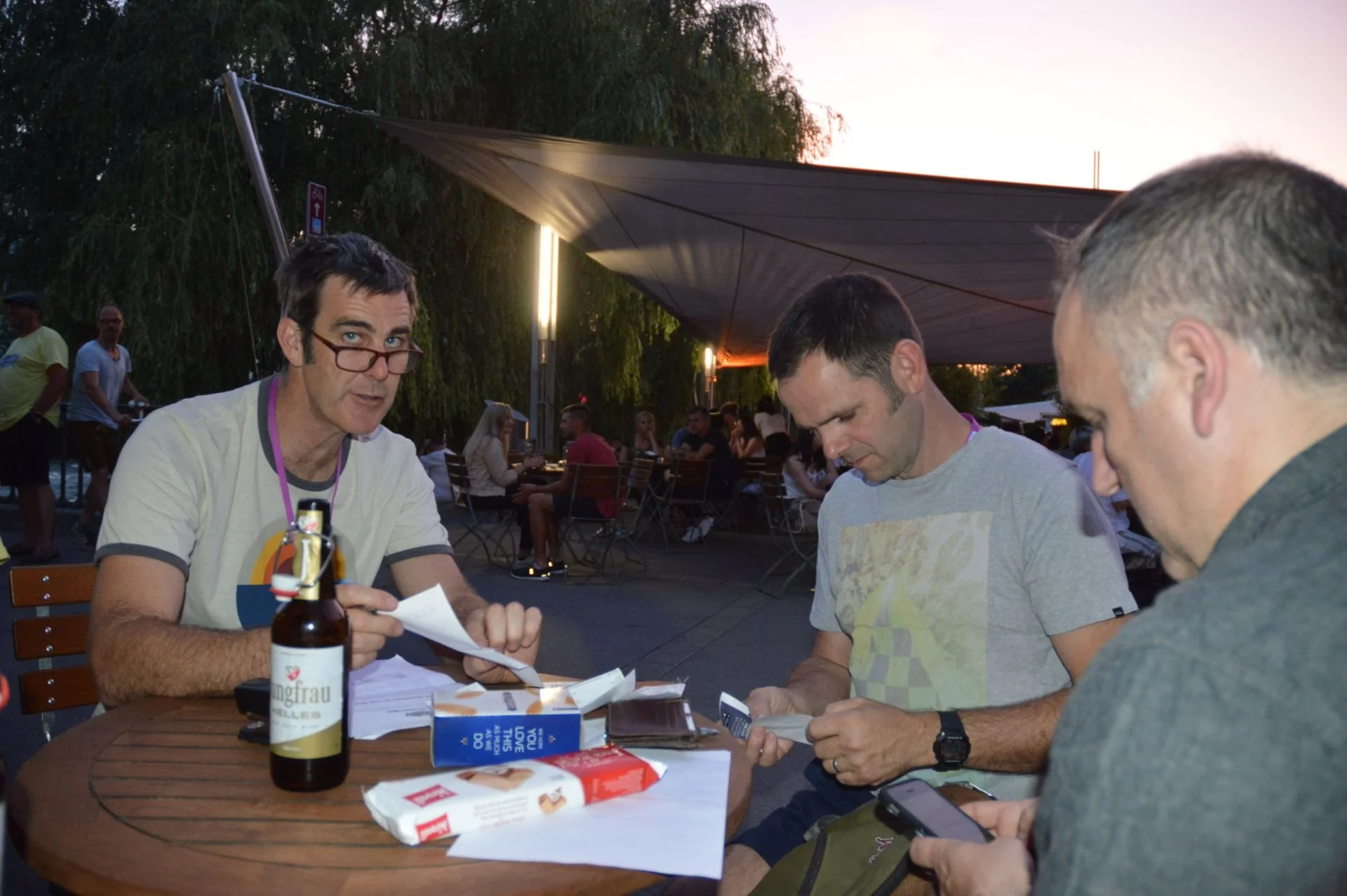 Three men sitting at a wooden table outdoors, engaged in conversation and looking at papers or mobile phones. One man has glasses and a beer bottle on the table, with other people in the background sitting under a canopy during twilight.