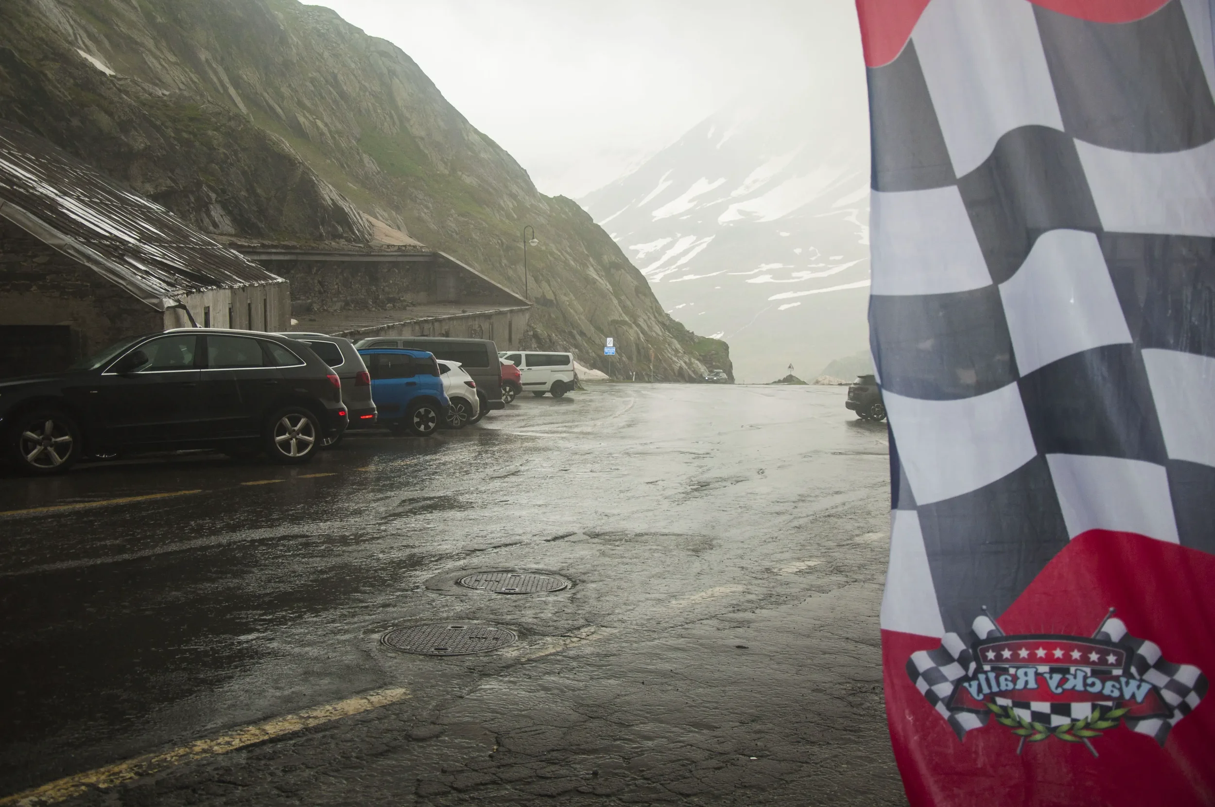 A mountain road with parked cars, a rocky hillside, and a checkered flag with a racing logo in the foreground during rainy weather.