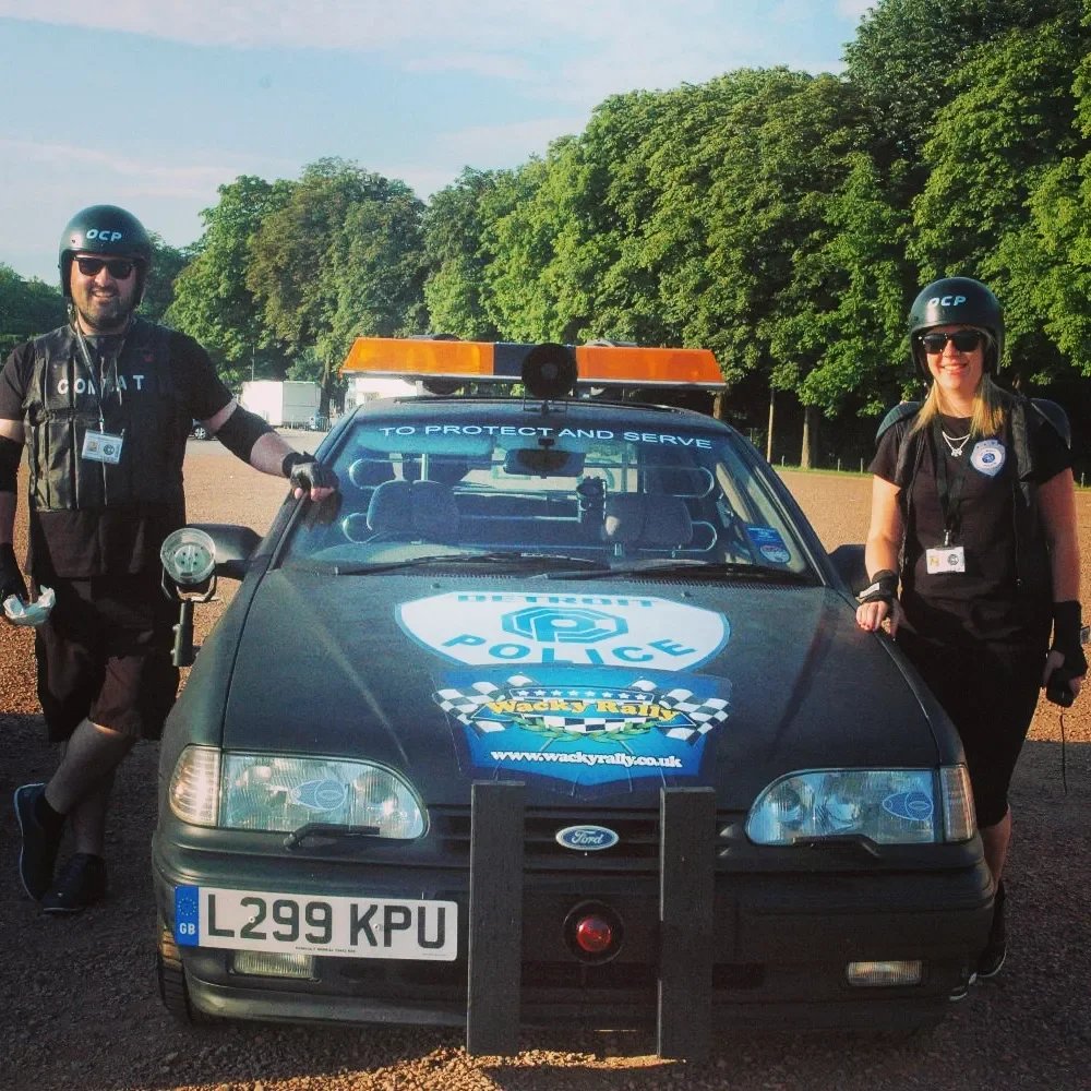 Two police officers in black uniforms and helmets stand beside a police car with a blue and white badge on the hood, in a park with green trees in the background.