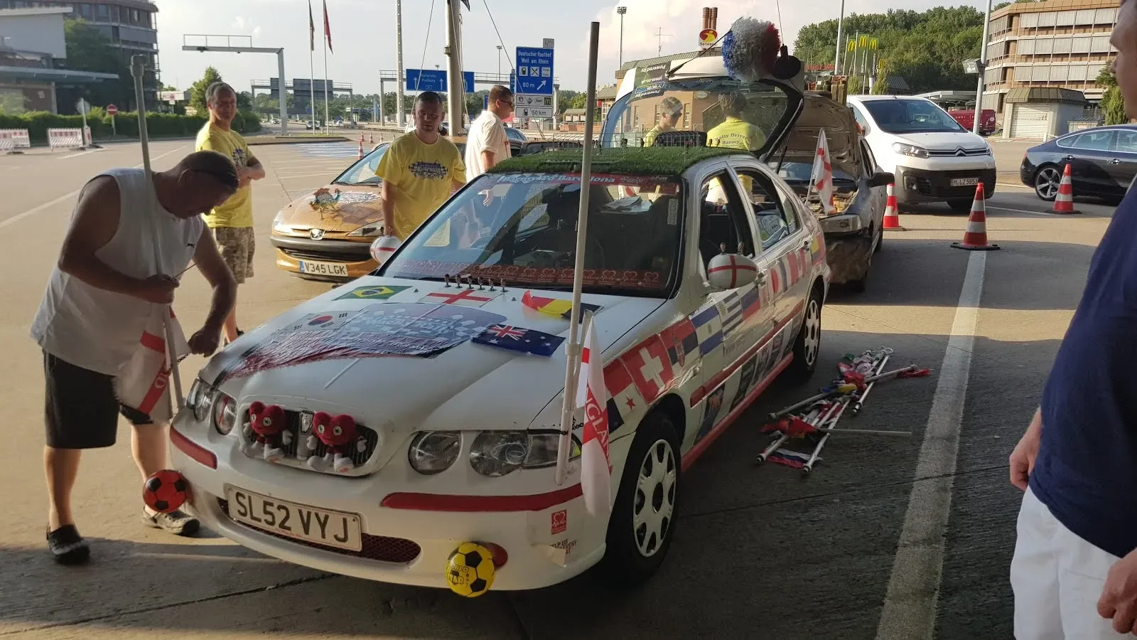 A white car decorated with various football-themed items and flags, parked on the street with people around it. The car has flags from different countries, plush toys, and soccer balls attached. People are interacting with the car, preparing or inspe