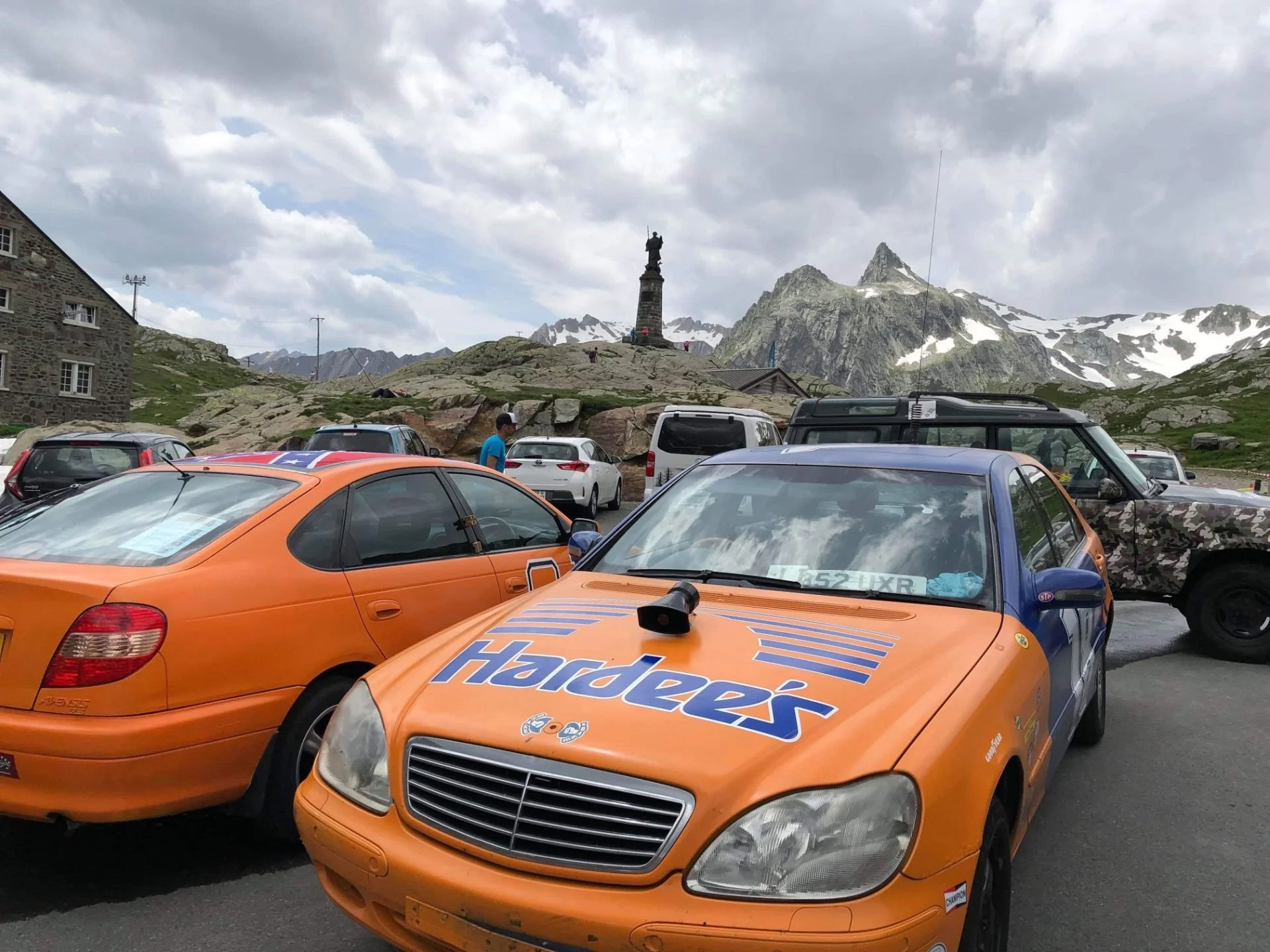 A parking lot with several cars, including a blue and orange car with Hardee's branding, surrounded by mountainous terrain with snow-capped peaks and a statue on a hill in the background.