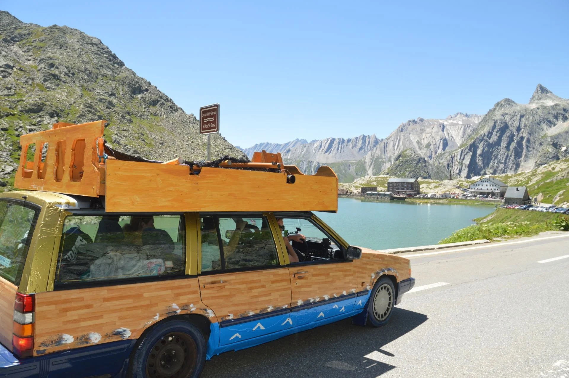 A custom-built car resembling a wooden house with a roof, driving along a mountain road near a lake, with mountains and buildings in the background.