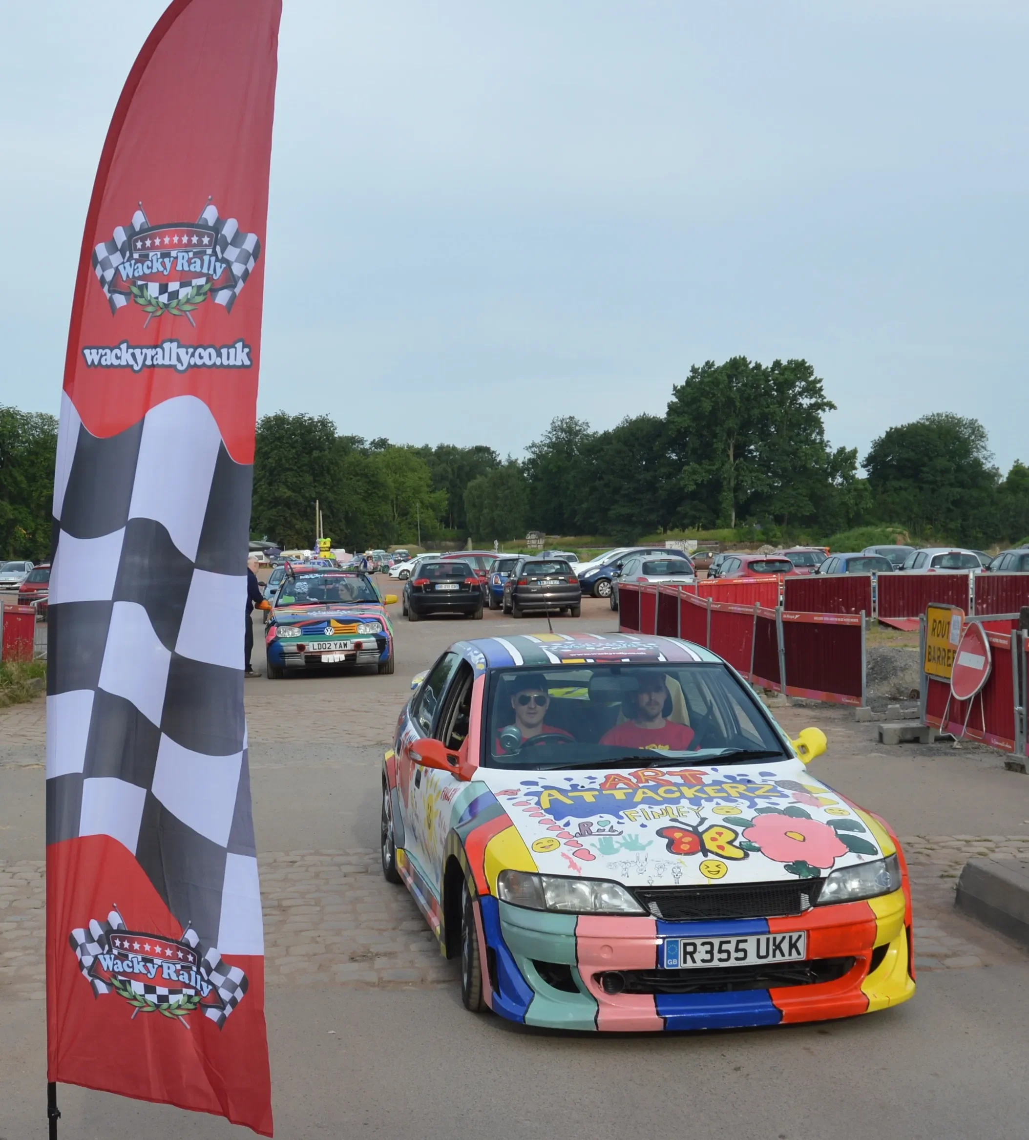 A colorful car with graffiti-style art on the hood and sides, displaying the words 'ART ATTACKERZ' and '8R', parked in front of a Wacky Rally flag near a parking lot with numerous cars and people.