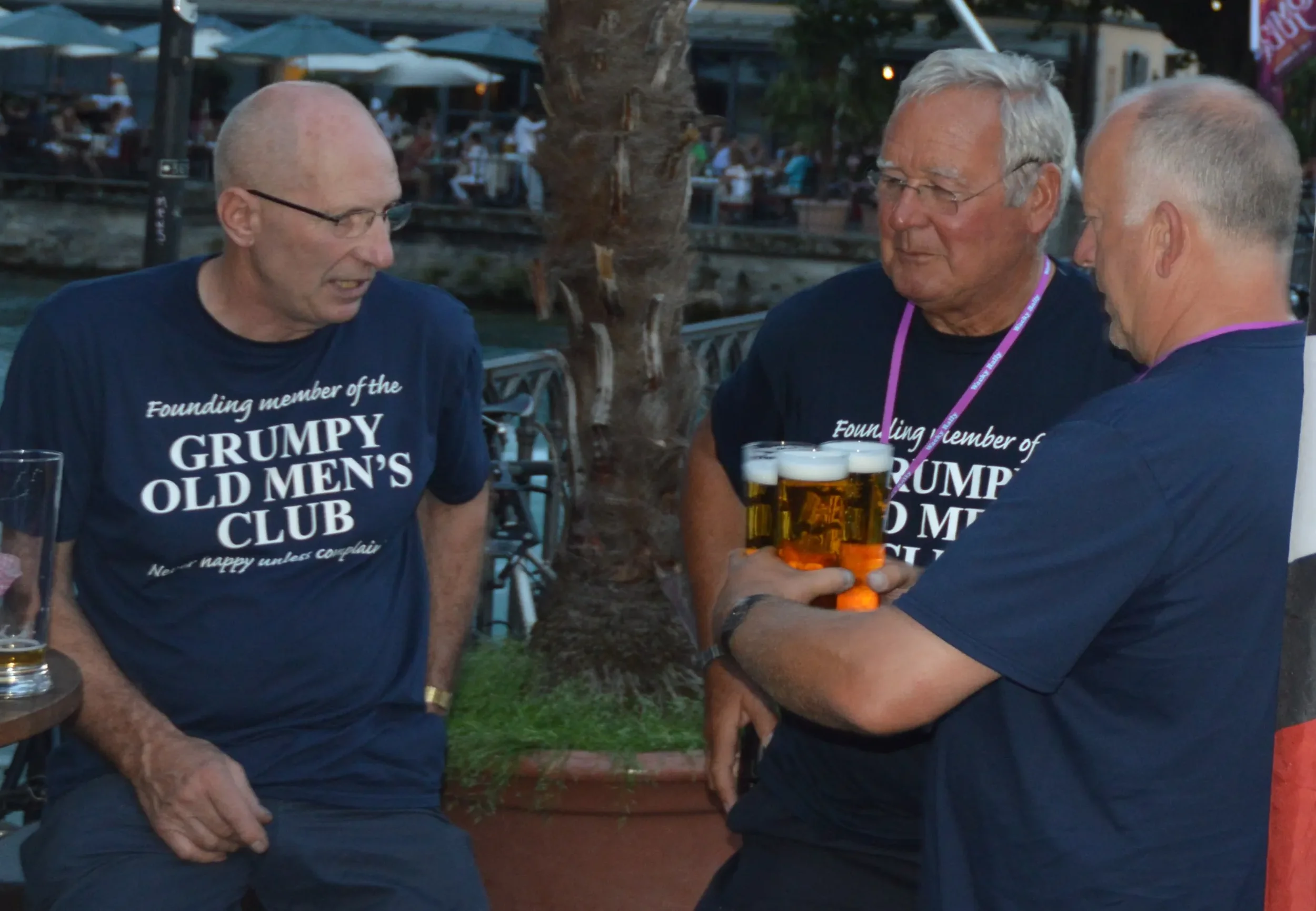 Three men wearing matching navy blue t-shirts with the text "Founding member of the Grumpy Old Men’s Club" are engaged in conversation. One man is holding four glasses of beer. They are outdoors in a social setting with other people and outdoor table