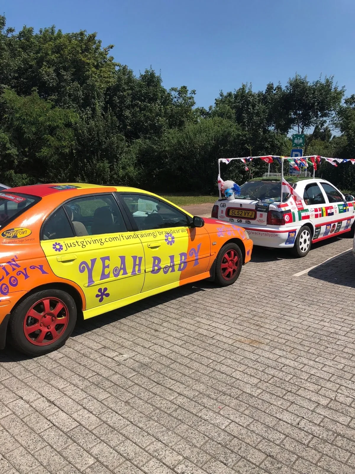 Two decorated cars parked side by side in a parking lot with trees in the background. The car in the foreground is yellow and orange with purple text that says 'YEAH BABY' and has the URL 'justgiving.com/fundraising/braudesry.' The car behind it is w