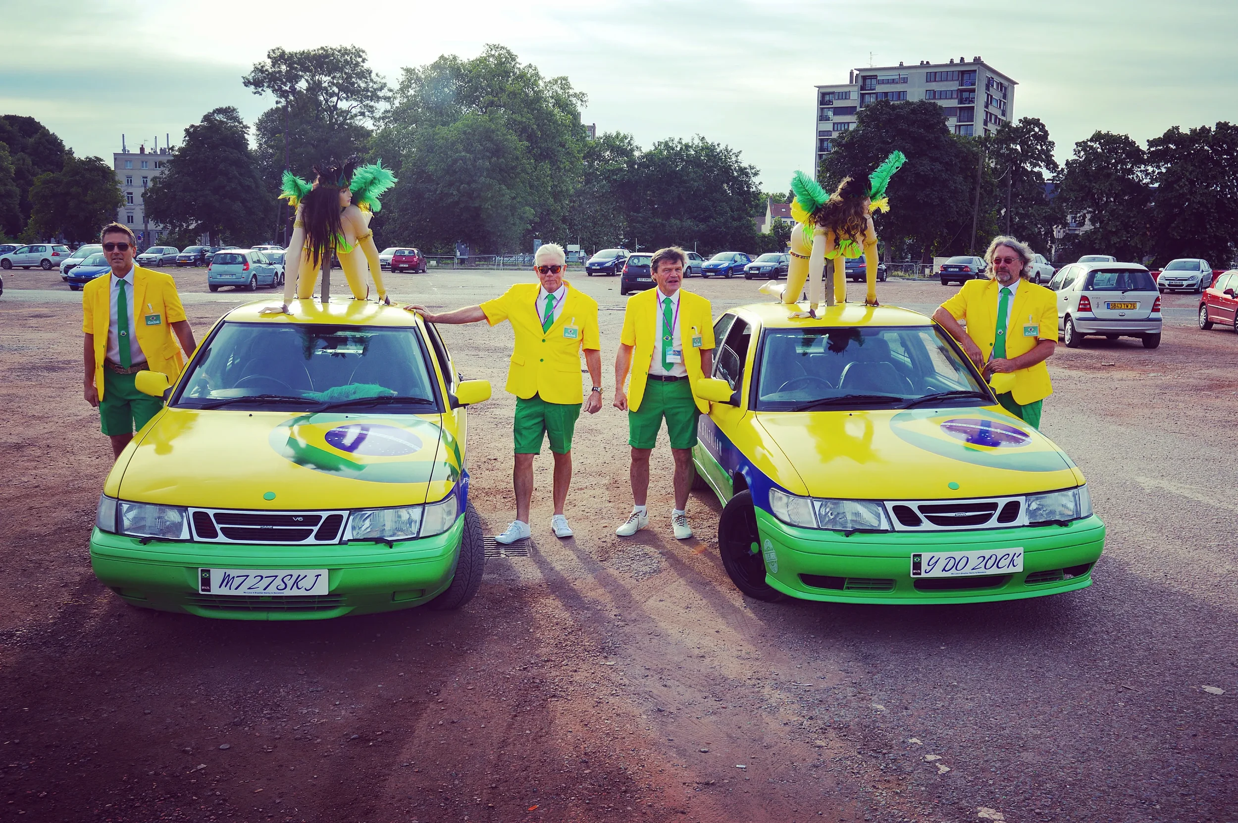 A group of six people dressed in yellow jackets, green pants, and green ties posing with two cars painted in bright yellow and green with logos and license plates, in an outdoor parking lot with trees, additional cars, and buildings in the background