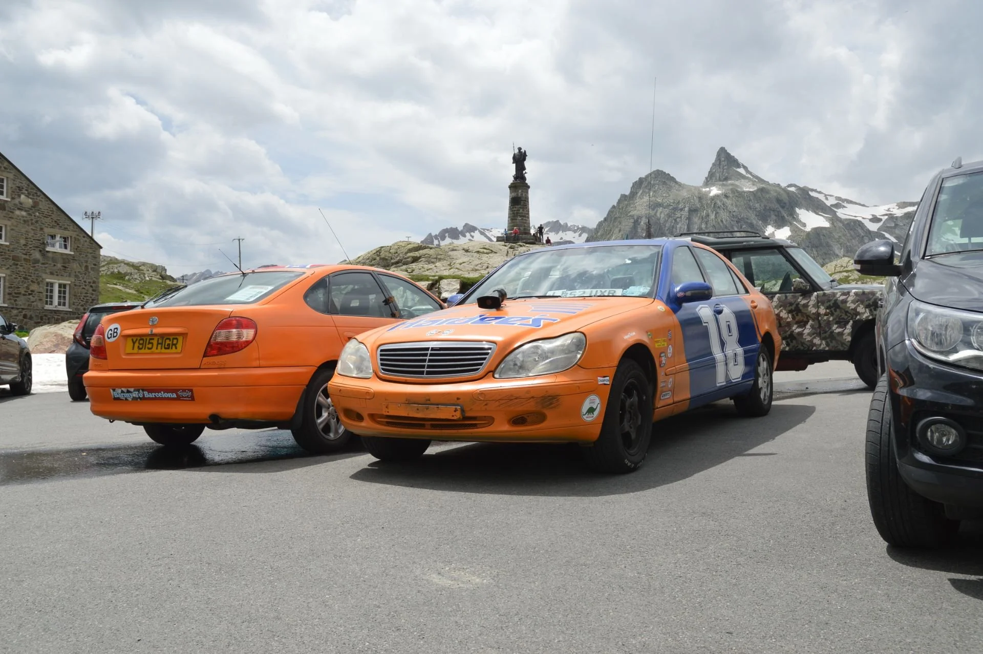 A parking lot with three cars, including a vintage orange Mercedes race car with the number 18, an orange Mazda coupe, and a black minivan. In the background, there are snow-capped mountains and a statue on a pedestal.