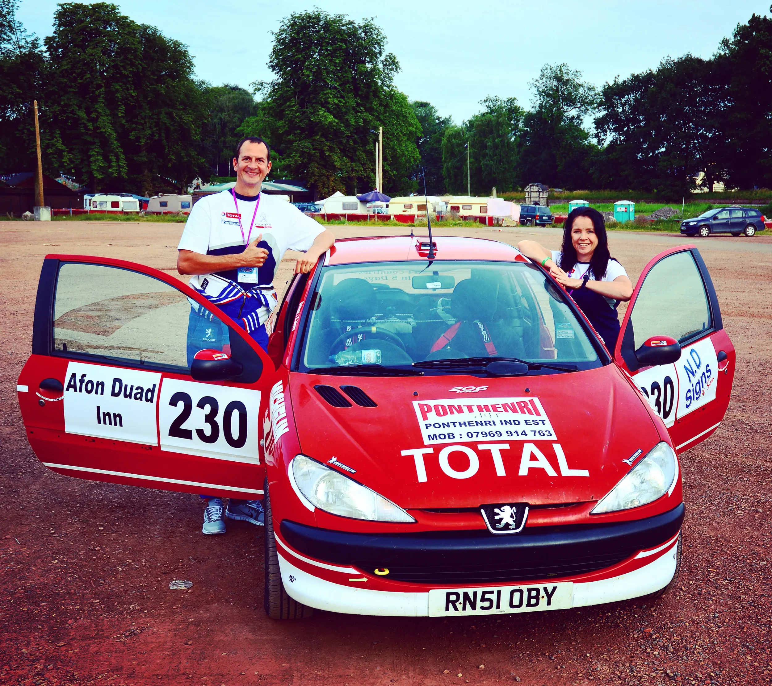 A man and woman standing beside a red rally car with racing numbers 230 and 30, parked on a dirt area with trees and parked vehicles in the background.